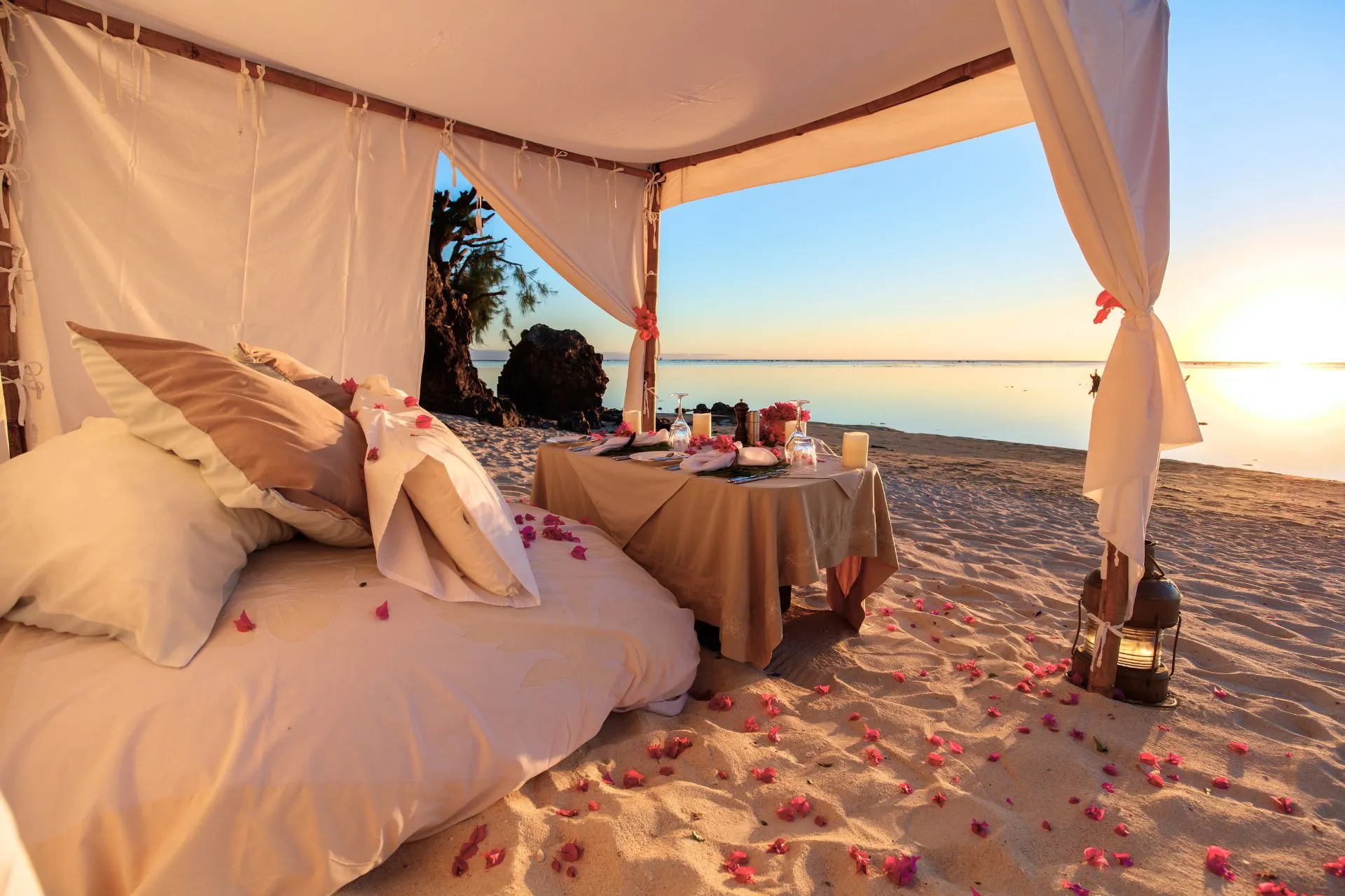 Een romantische setting op het strand met een bed bedekt met rozenblaadjes, terwijl de zonsondergang de lucht in warme kleuren schildert. Het perfecte intieme moment, omringd door de rust van de oceaan.