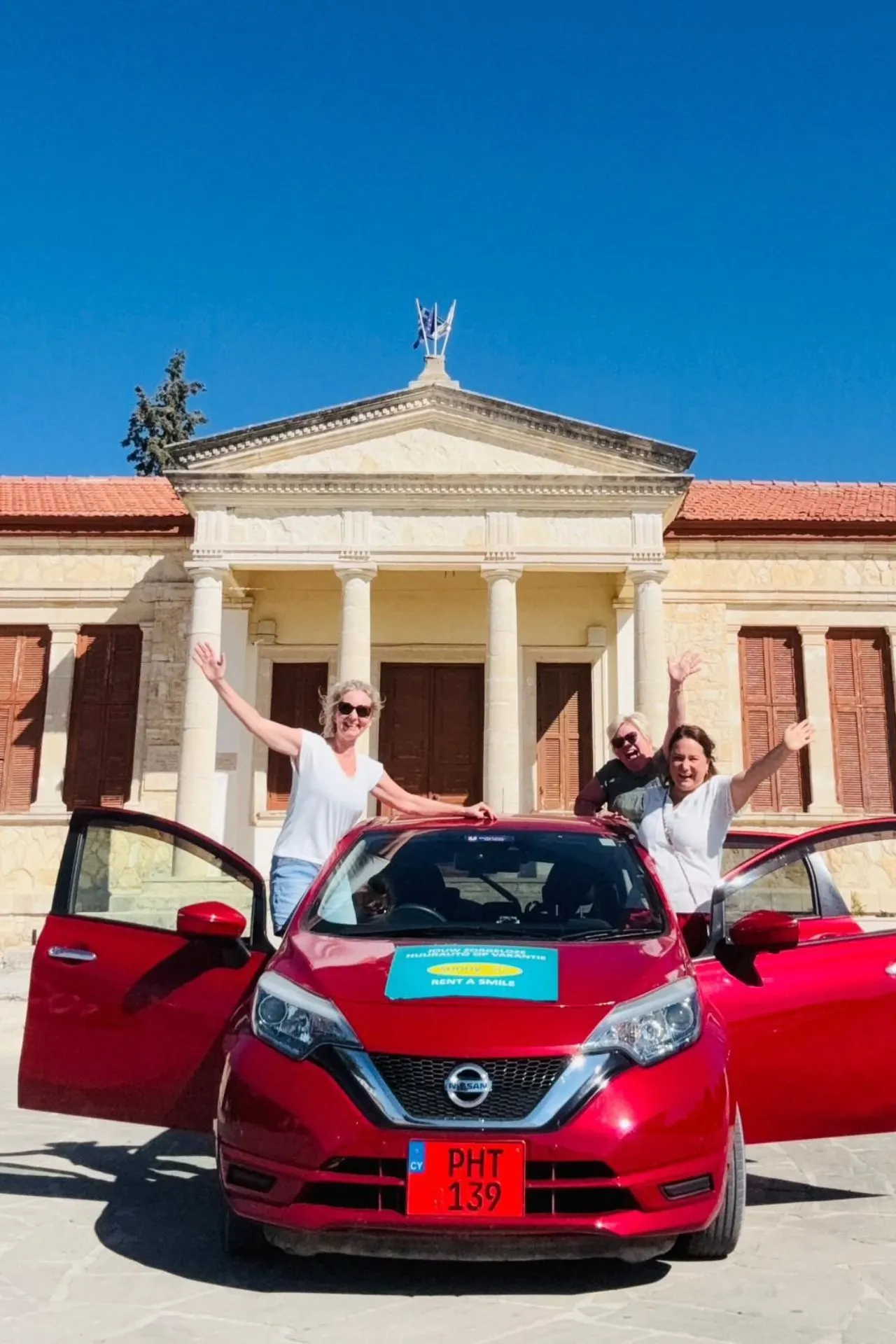 Rode huurauto van Sunny Cars geparkeerd voor een klassiek gebouw met zuilen en houten luiken in Cyprus. Drie personen staan bij de auto met open portieren.