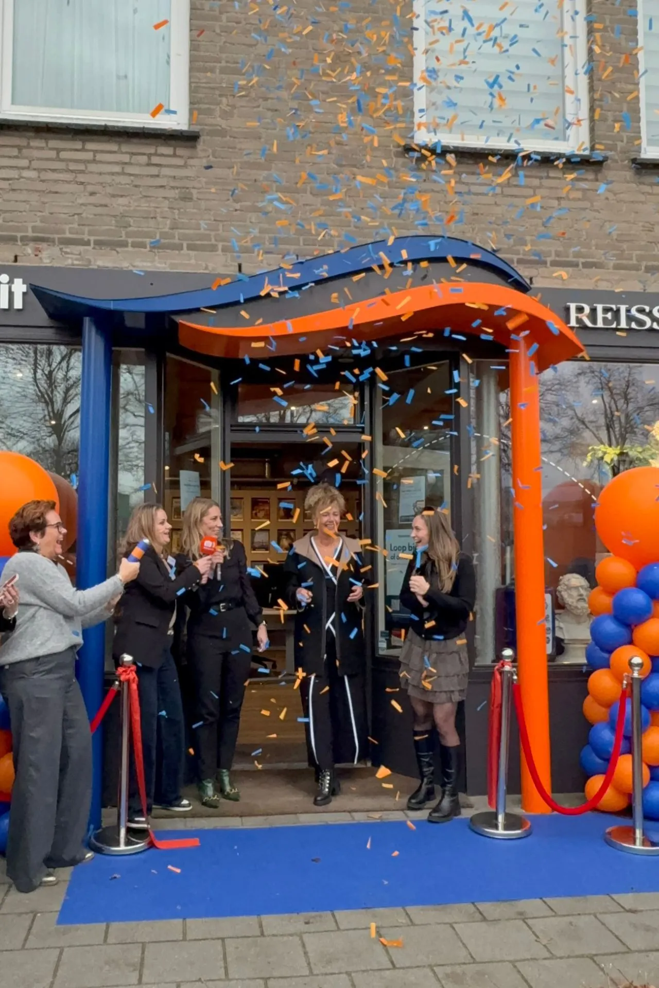 Officieel openingsmoment van Travelcenter de Wit Asten met oranje en blauwe confetti bij de entree, omringd door feestelijke ballonnen en een blauwe loper.