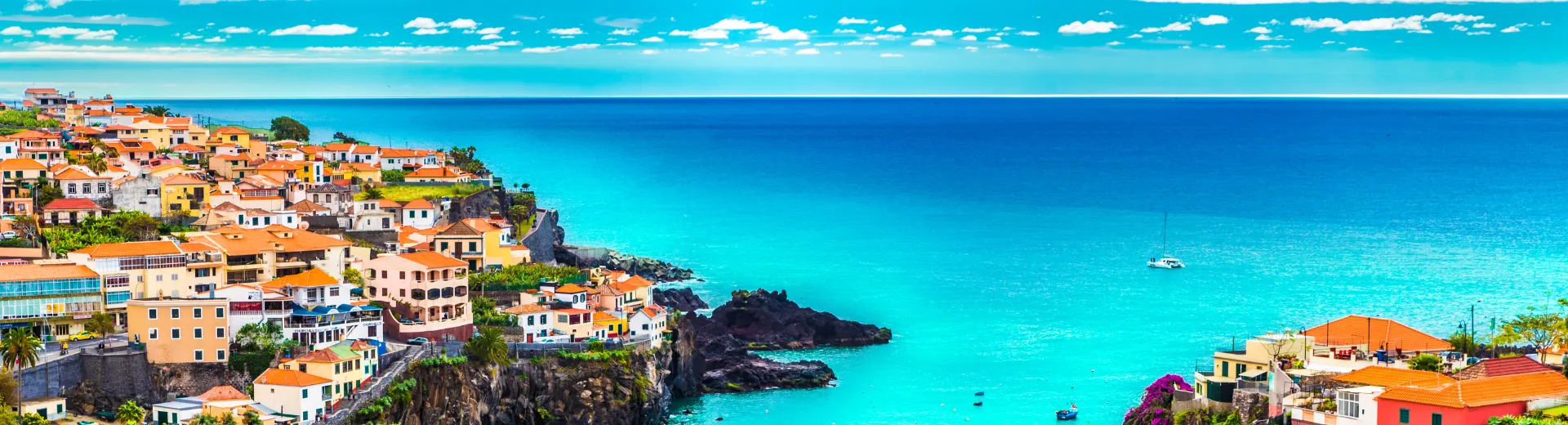 Schitterend uitzicht op een kustplaats met kleurrijke huizen en rode daken die tegen een groene heuvel liggen, uitkijkend over helder turquoise water van de Atlantische Oceaan. Donkere rotsformaties langs de kust zorgen voor een indrukwekkend contrast met het azuurblauwe water. Aan de horizon strekt de oceaan zich eindeloos uit onder een heldere lucht met witte wolken. Perfecte bestemming voor een zonvakantie, wandelingen langs kliffen en het ontdekken van authentieke mediterrane charme.