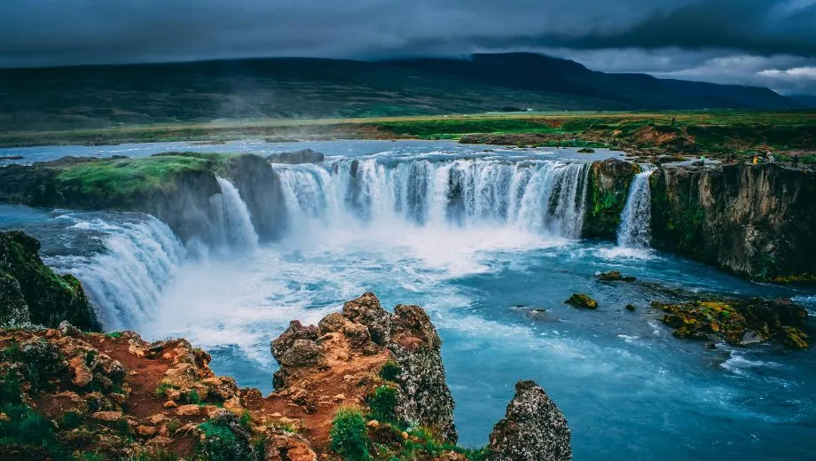 Indrukwekkende Goðafoss-waterval in IJsland, waar breed water naar beneden stort in een blauwe rivier, omgeven door ruige rotsen en groene vlaktes.