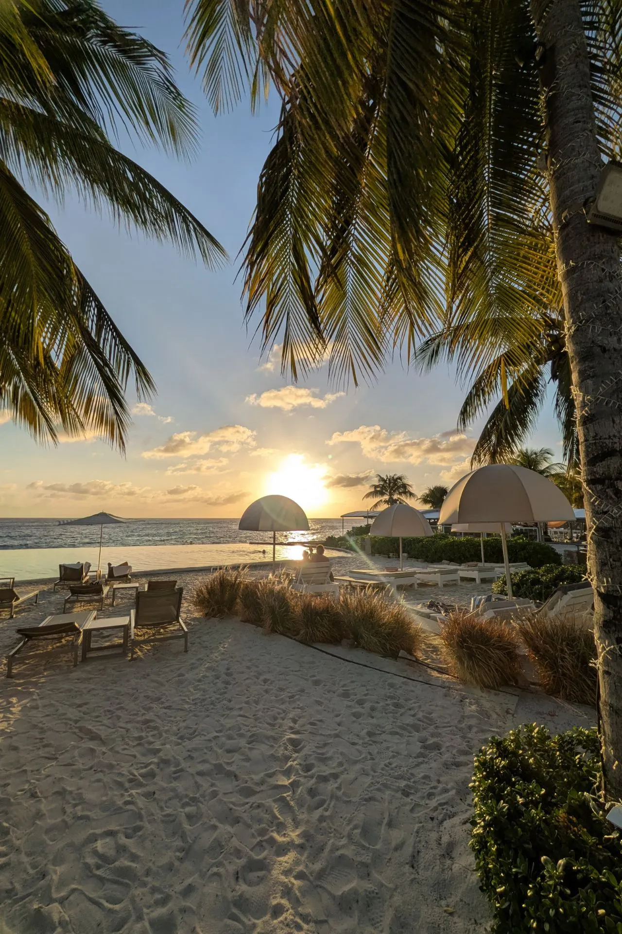 Adembenemende zonsondergang op een wit zandstrand met palmbomen en ligbedden op Curaçao. De zon reflecteert op de rustige zee, terwijl het strand een serene sfeer biedt voor ontspanning. Ideaal voor romantische vakanties, luxe resorts en genieten van het Caribische klimaat.