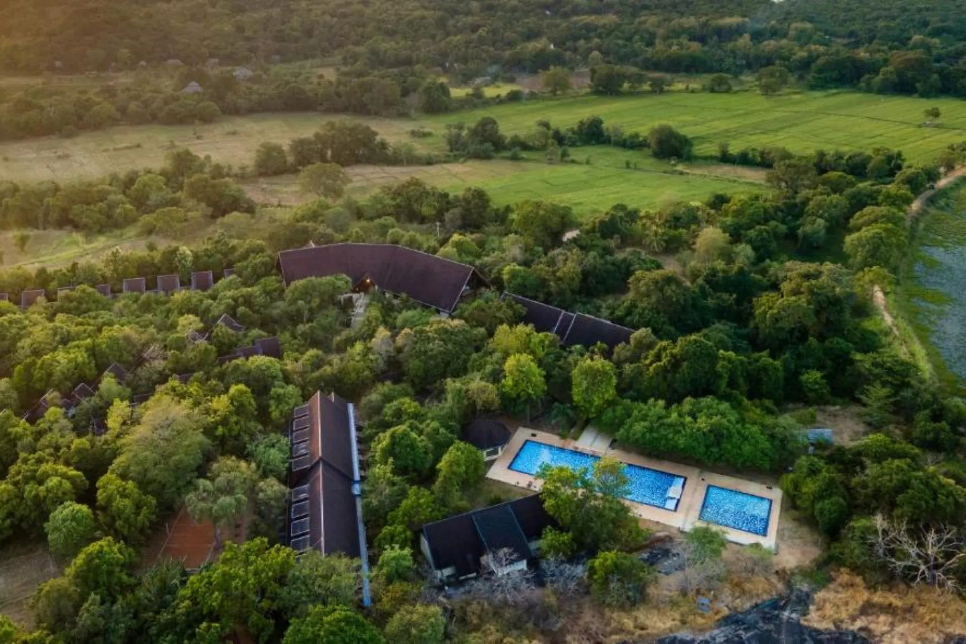 Luchtfoto van Occidental Paradise Dambulla in Sri Lanka, midden in het groen met verspreide villa’s, twee zwembaden en uitzicht op rijstvelden en heuvelachtig landschap bij zonsondergang.