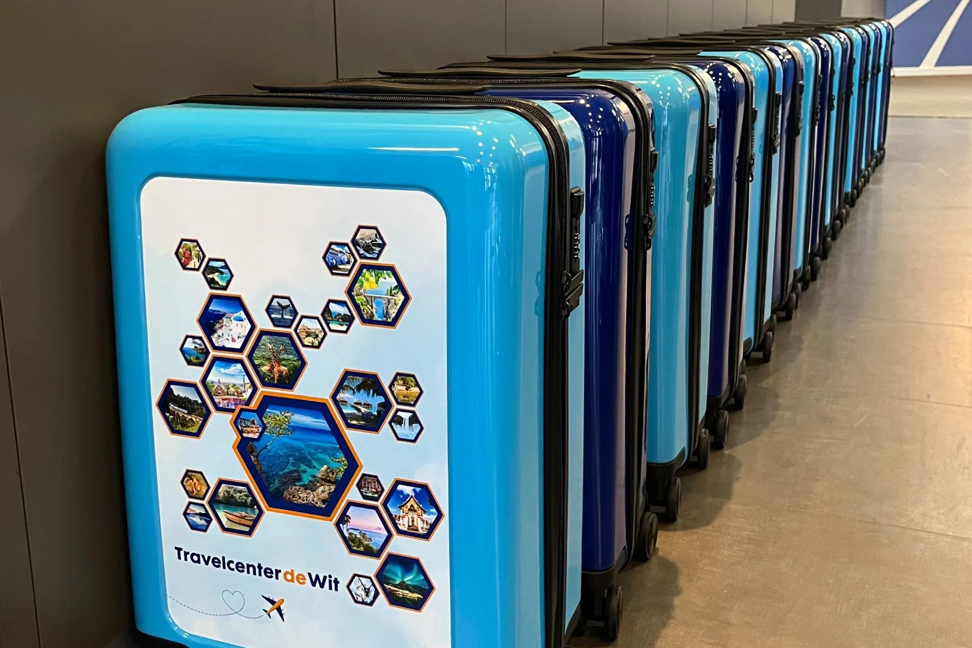 Een rij felblauwe en donkerblauwe koffers met een bedrukte voorkant, waarop diverse reisbestemmingen in zeshoekige frames te zien zijn. De koffers zijn onderdeel van een promotieactie van Travelcenter de Wit en staan op een rij in een moderne, strakke omgeving.