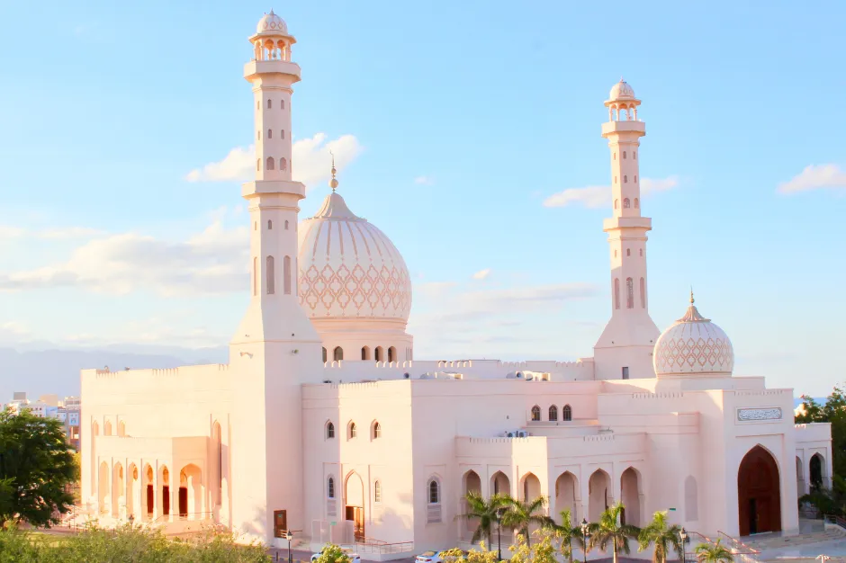 Prachtige witte moskee in Oman met twee hoge minaretten en een grote centrale koepel, omringd door groene palmbomen en gelegen tegen een achtergrond van blauwe lucht en lichte wolken. De architectuur straalt elegantie en rust uit, met verfijnde details en een imposante entree. Deze foto benadrukt de culturele rijkdom en religieuze tradities van Oman, ideaal voor reizigers die op zoek zijn naar authentieke bezienswaardigheden en indrukwekkende architectuur tijdens hun reis.