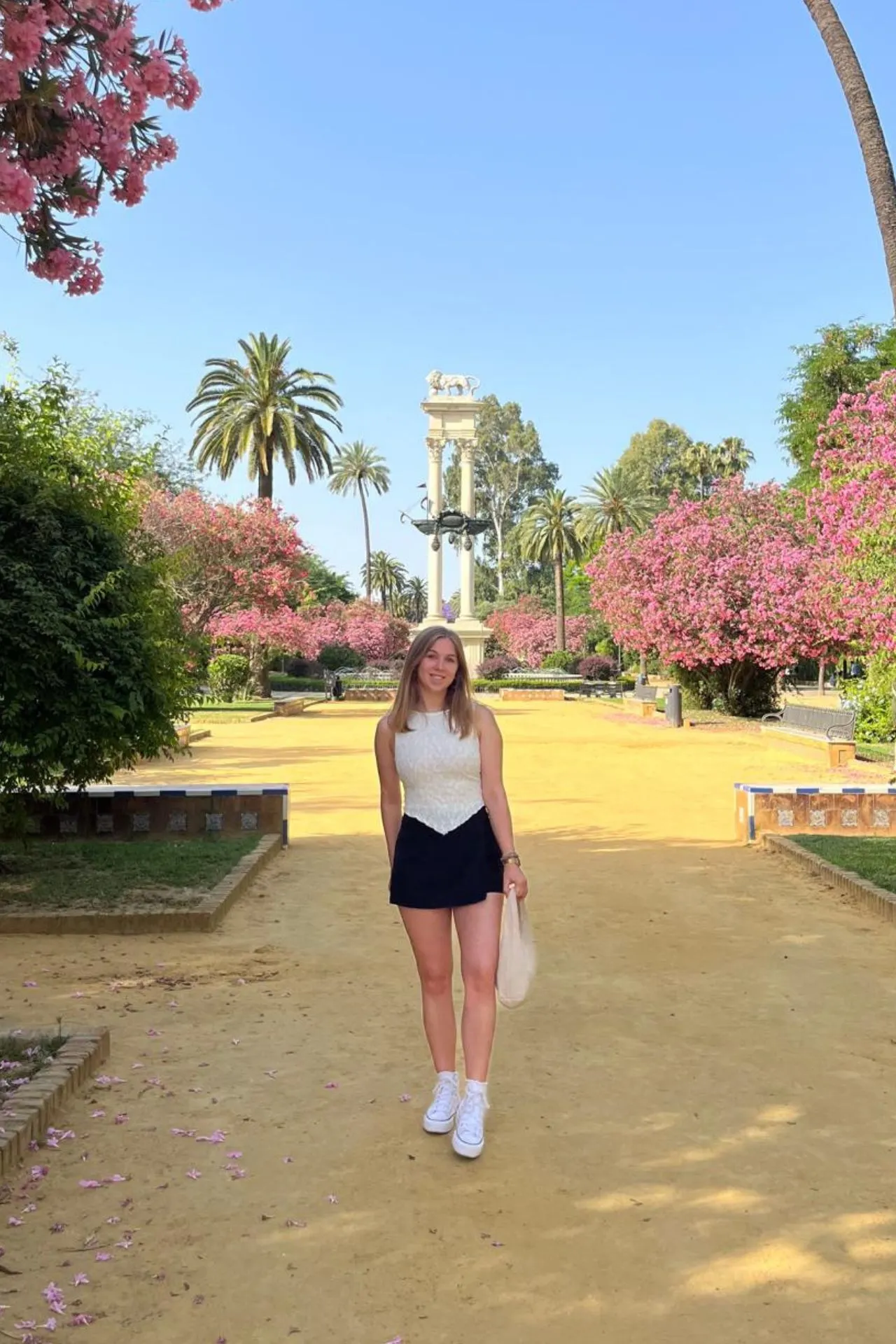 Marketeer Martje poseert glimlachend op een zonovergoten wandelpad in het María Luisa Park in Sevilla, omringd door palmbomen en bloeiende roze oleanders. Op de achtergrond staat het monument van Columbus met twee witte zuilen en een schip erop.