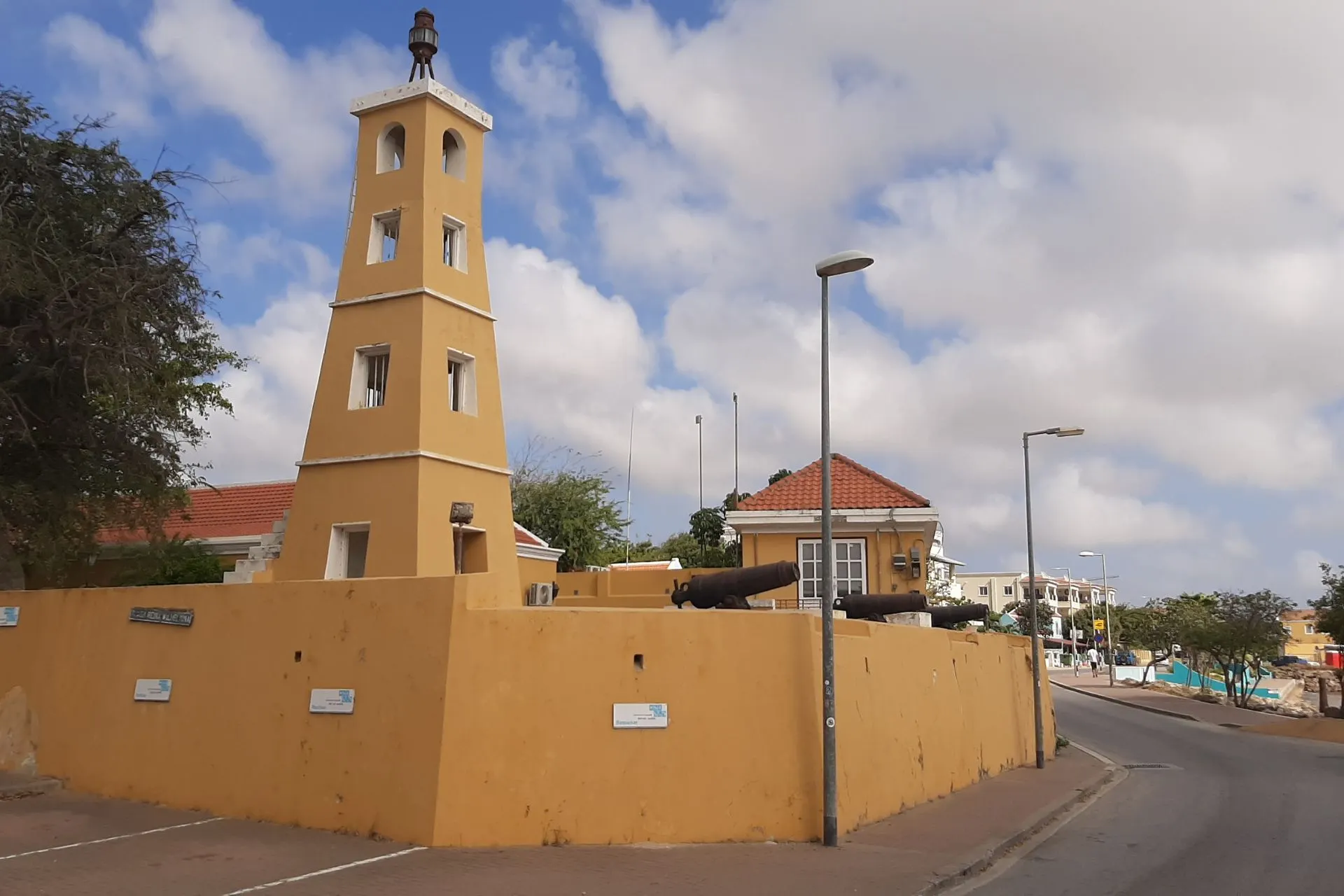 Historische gele forttoren in Kralendijk, Bonaire, omringd door een stevige muur en oude kanonnen. Dit iconische gebouw weerspiegelt de koloniale architectuur en het rijke culturele erfgoed van het eiland. Bonaire is niet alleen beroemd om zijn stranden en duikspots, maar ook om zijn monumenten en geschiedenis. De toren staat in een charmante straat met traditionele huizen en blauwe lucht, ideaal voor sightseeing en het ontdekken van de authentieke sfeer van Bonaire.