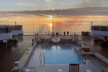 Een panoramisch uitzicht vanaf het buitendek van de Nieuw Statendam van Holland America Line, met een zwembad en jacuzzi's op de voorgrond en een prachtige zonsondergang boven de oceaan