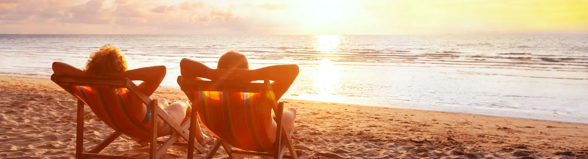 Een serene afbeelding van twee mensen in strandstoelen, ontspannen achteroverleunend met hun handen achter hun hoofd, terwijl ze genieten van een prachtige zonsondergang aan de horizon. Het beeld straalt zorgeloosheid en rust uit, perfect passend bij het thema van onbezorgd op reis.