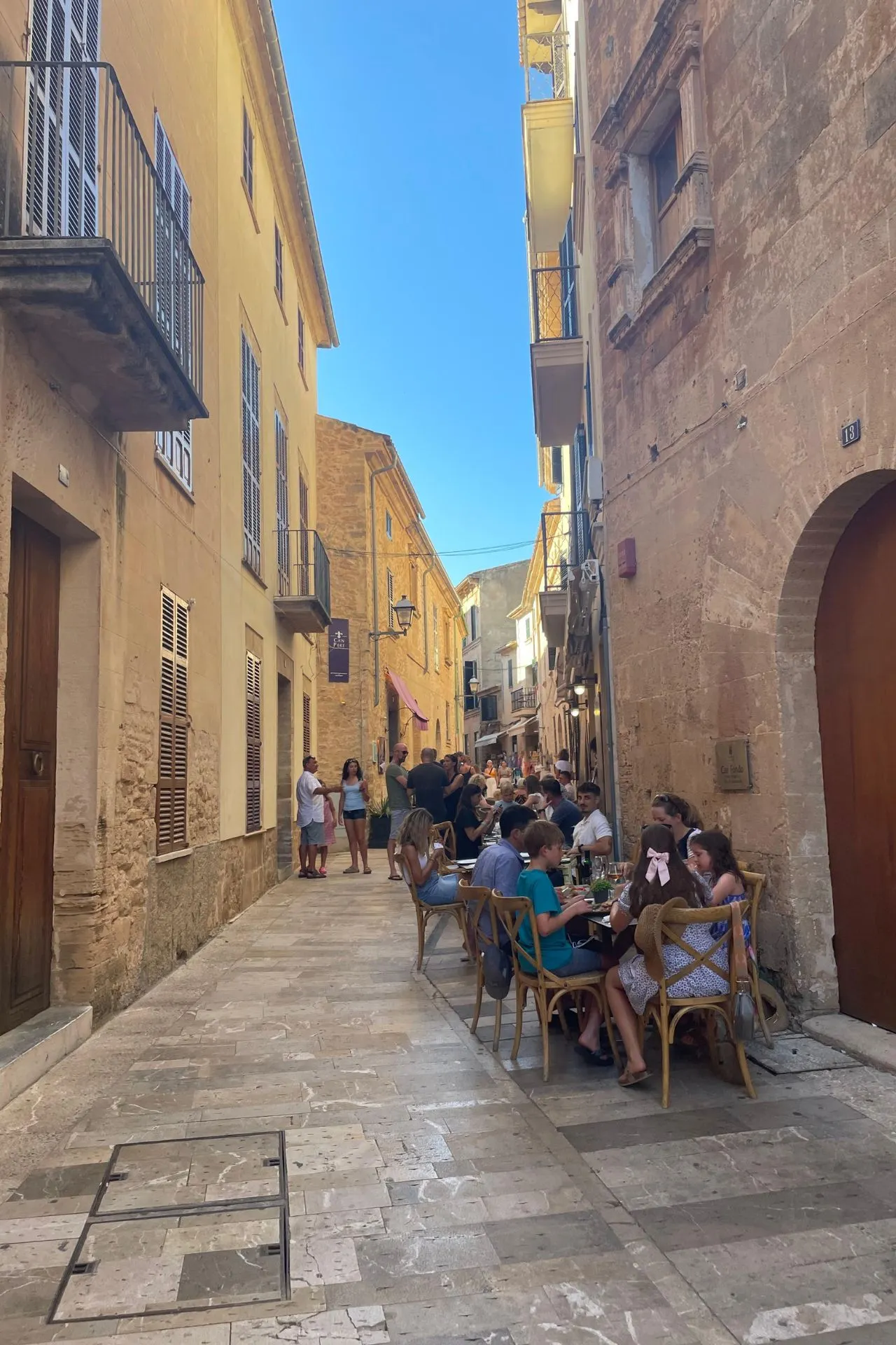 Mensen zitten aan tafeltjes langs een smalle geplaveide straat tussen oude zandkleurige gebouwen in Alcúdia, onder een heldere blauwe lucht.
