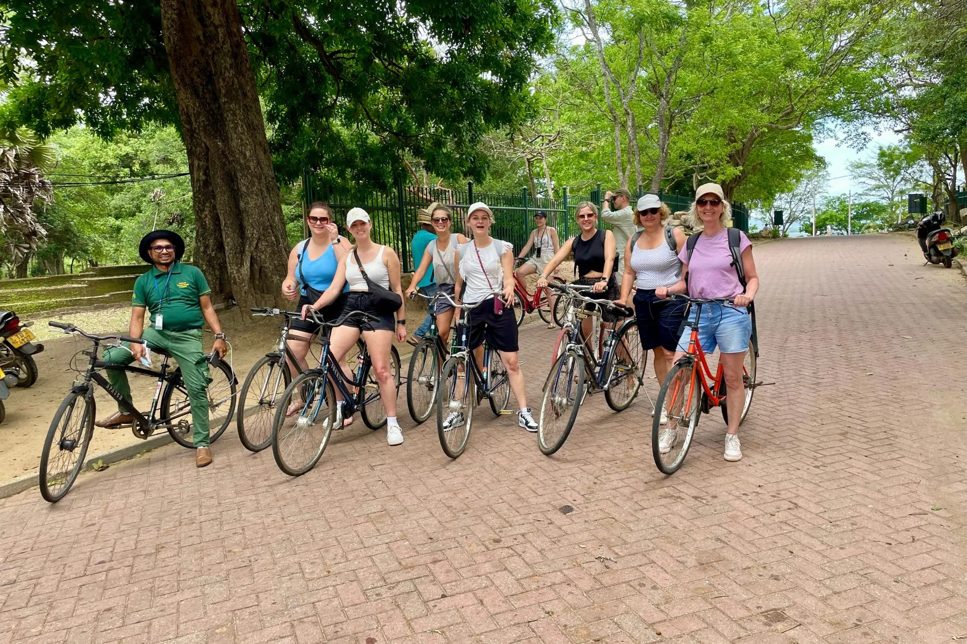 Groep vrouwen op fietsen, glimlachend en klaar voor vertrek, tijdens een fietstocht door de historische ruïnes van Polonnaruwa. De schaduwrijke weg wordt omzoomd door een ijzeren hek en dichte bomen, wat zorgt voor een aangename, ontspannen sfeer terwijl ze het UNESCO-werelderfgoed verkennen op een sportieve manier.