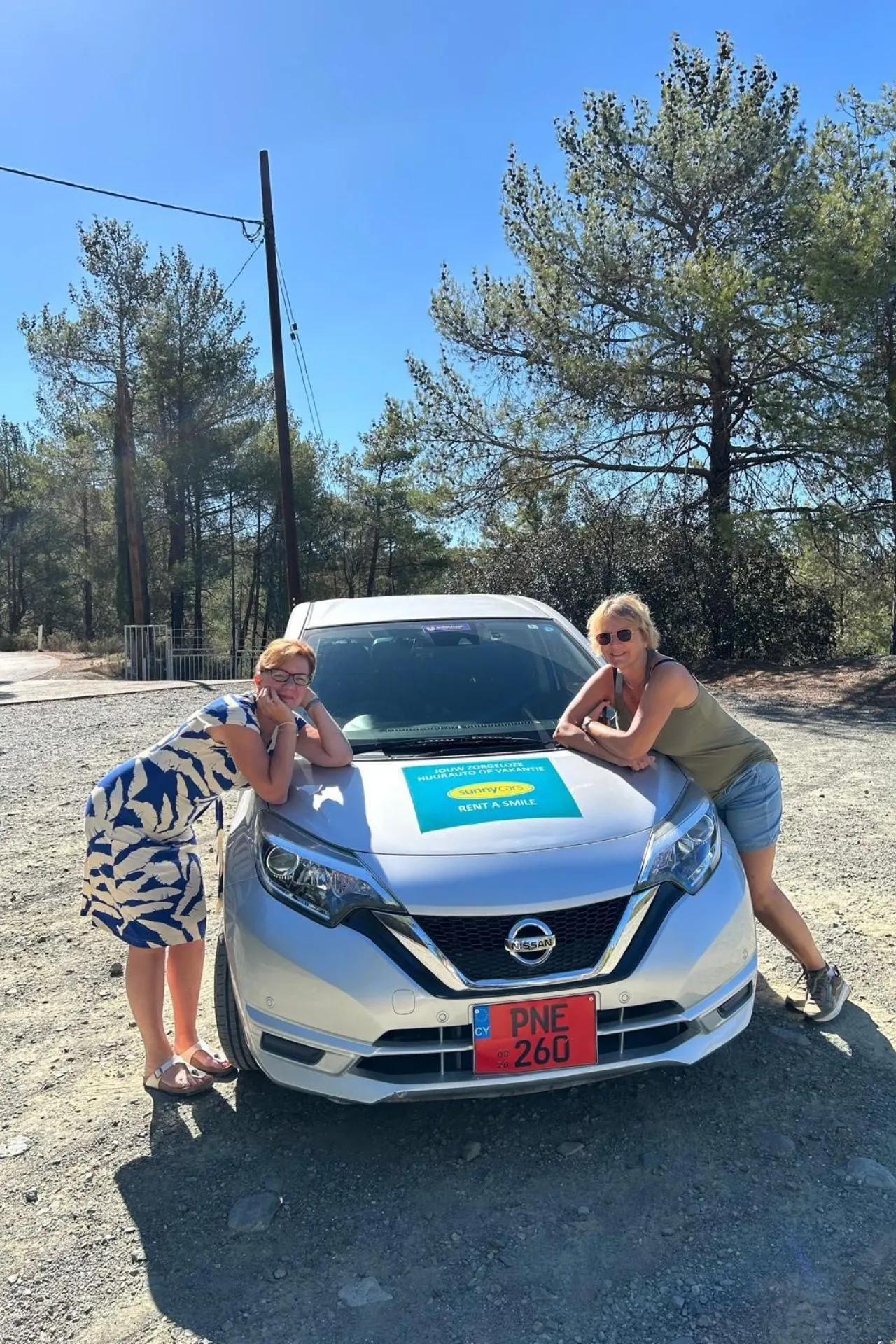 Witte huurauto van Sunny Cars op een onverharde weg in Cyprus, omringd door bomen. Twee personen leunen op de motorkap.