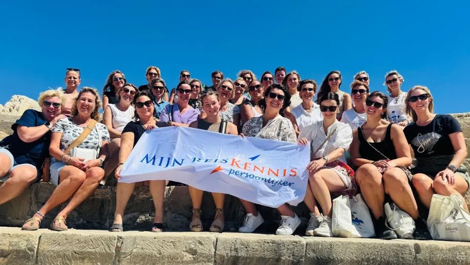 
Groepsfoto van mensen die samen op stenen tribunes zitten bij een archeologische site, met een helderblauwe lucht op de achtergrond. Voor hen wordt een witte banner met het logo en de tekst “Mijn ReisKennis Persoonlijker” vastgehouden.