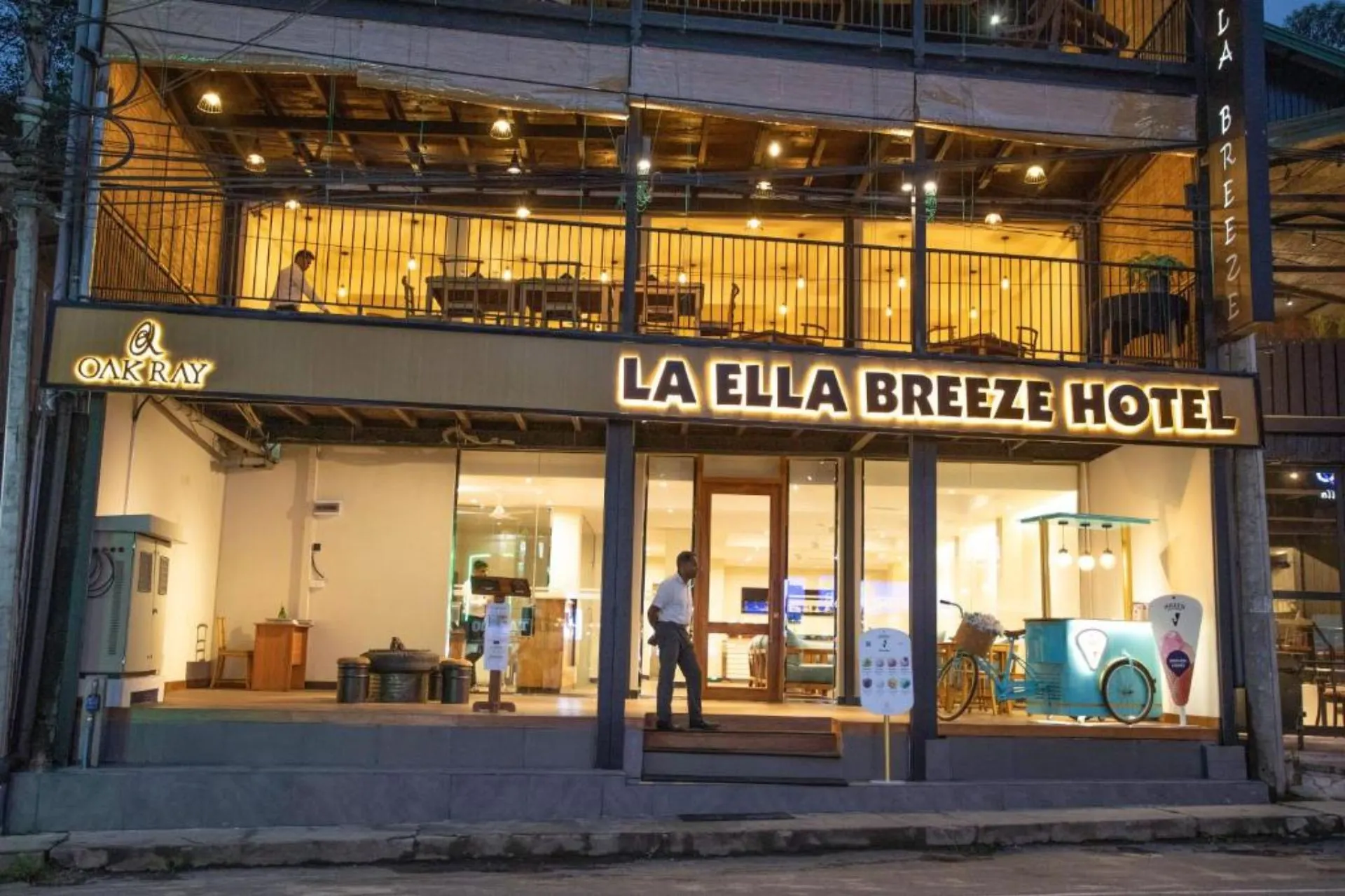 Avondaanzicht van Oak Ray La Ella Breeze Hotel in Ella met verlichte gevel, glazen entree, bovenliggend restaurant met balkon en een opvallende ijswagen in de lobby.