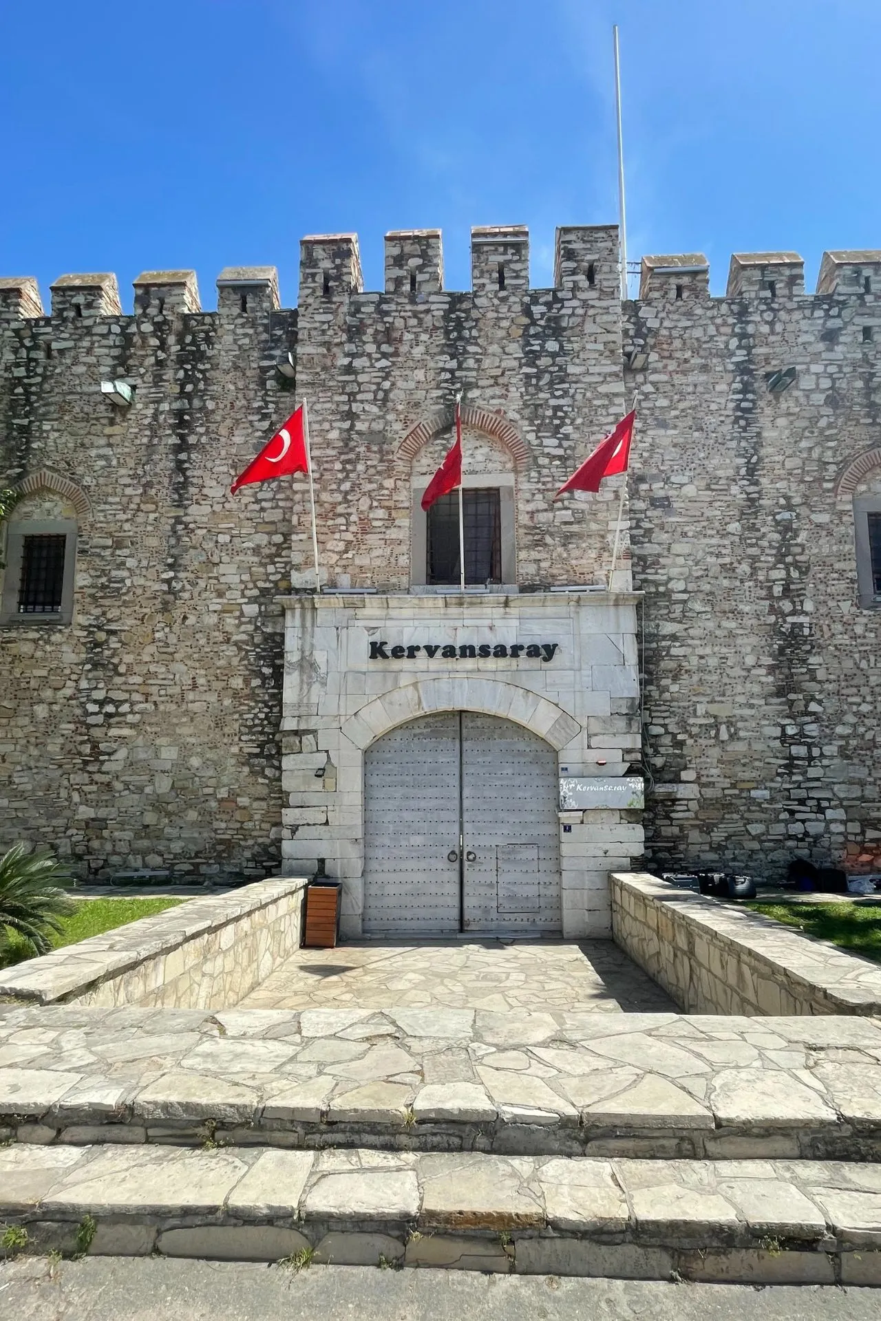 Vooraanzicht van het historische Kervansaray in Kuşadası, met Turkse vlaggen en een robuuste stenen gevel.