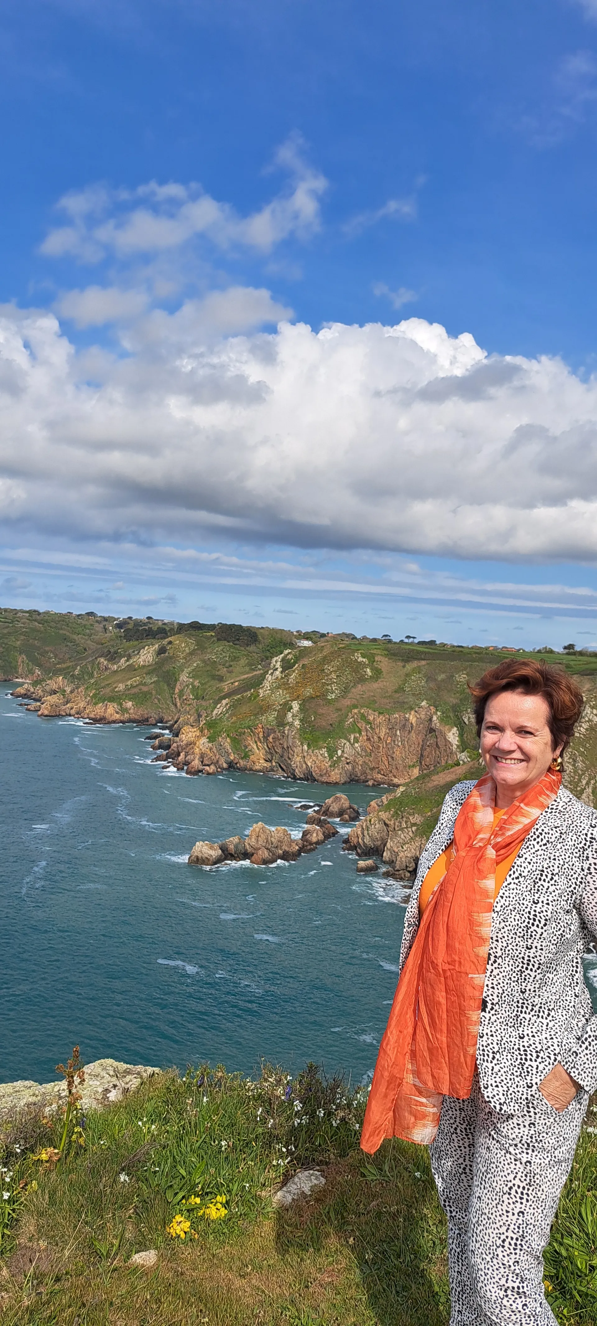 Spectaculair uitzicht vanaf een klif met helderblauw zeewater en ruige rotsformaties langs de kustlijn, gefotografeerd onder een stralende blauwe lucht met enkele wolken. Op de voorgrond staat een persoon in een lichtgrijze outfit met een opvallende oranje sjaal, omringd door groene vegetatie en gele bloemen. Perfect voor natuurwandelingen, kustfotografie en avontuurlijke vakanties met panoramische uitzichten op zee en rotsachtige landschappen.