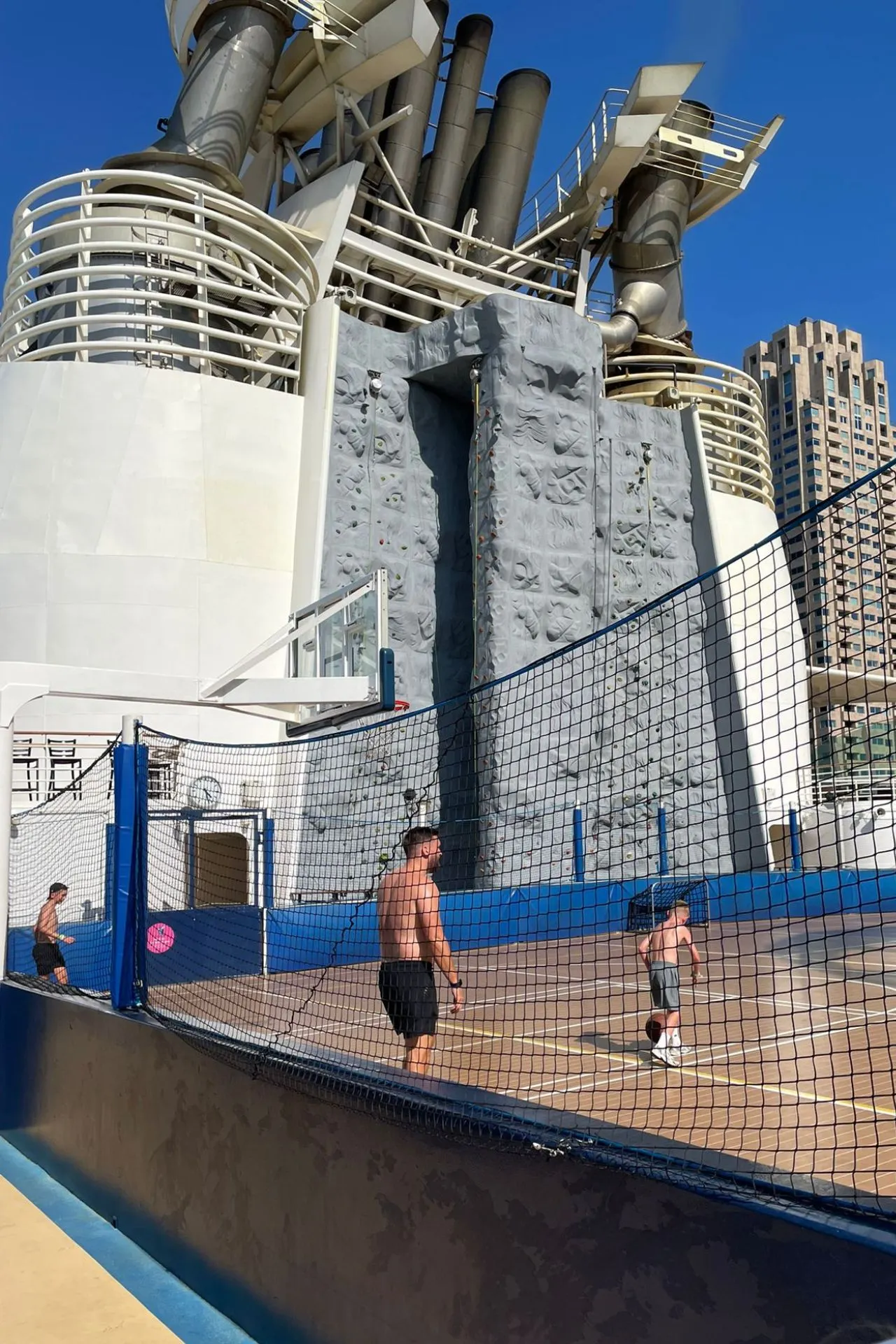 Sportveld op het dek van de Independence of the Seas met basketbalspelers, op de achtergrond een hoge klimwand tegen de schoorsteenconstructie van het schip.