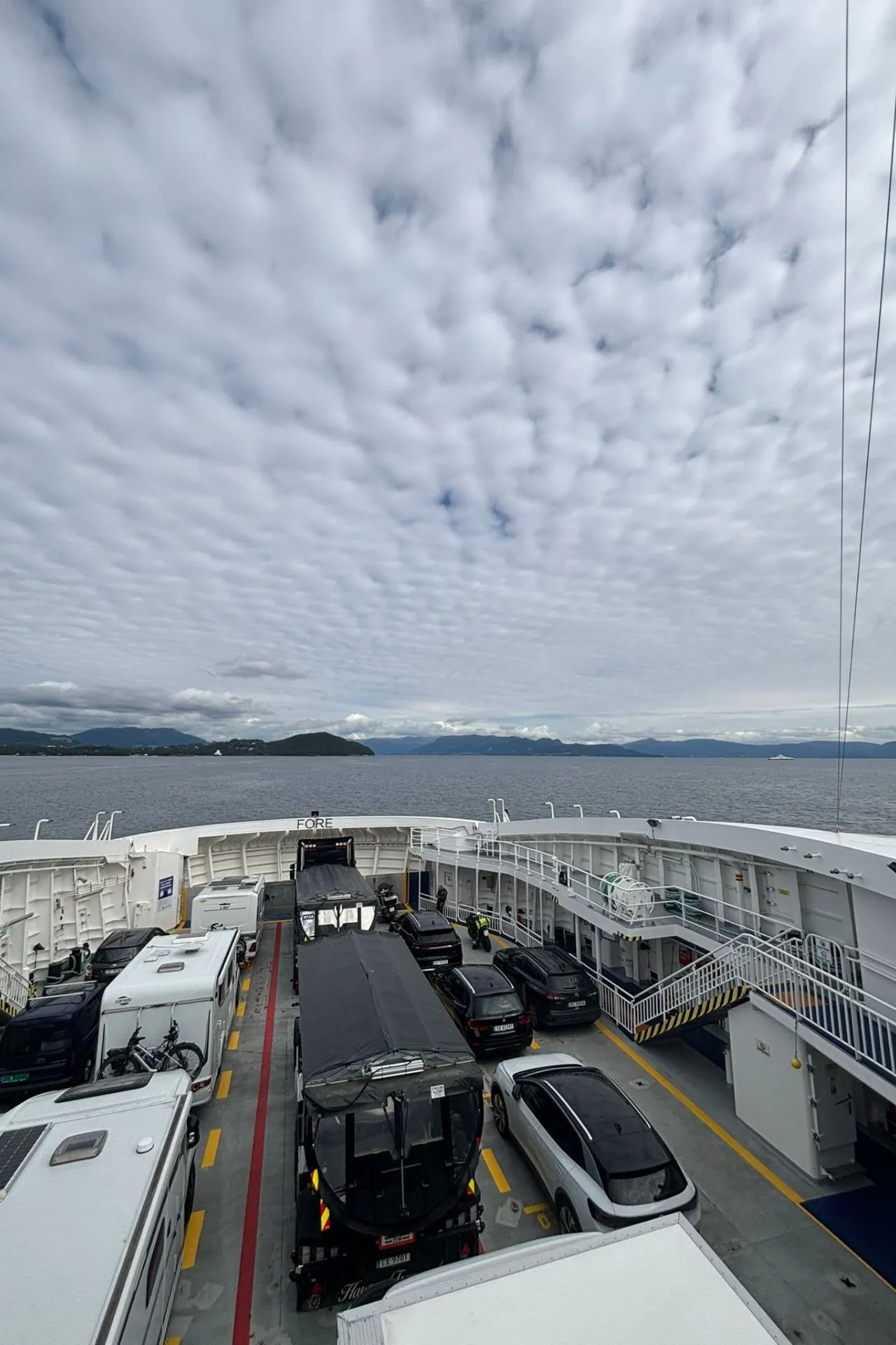 Voertuigen geparkeerd op het open dek van een veerboot, waaronder campers, auto's en een vrachtwagen. Op de achtergrond is een uitgestrekt wateroppervlak zichtbaar met bergen aan de horizon onder een bewolkte lucht.