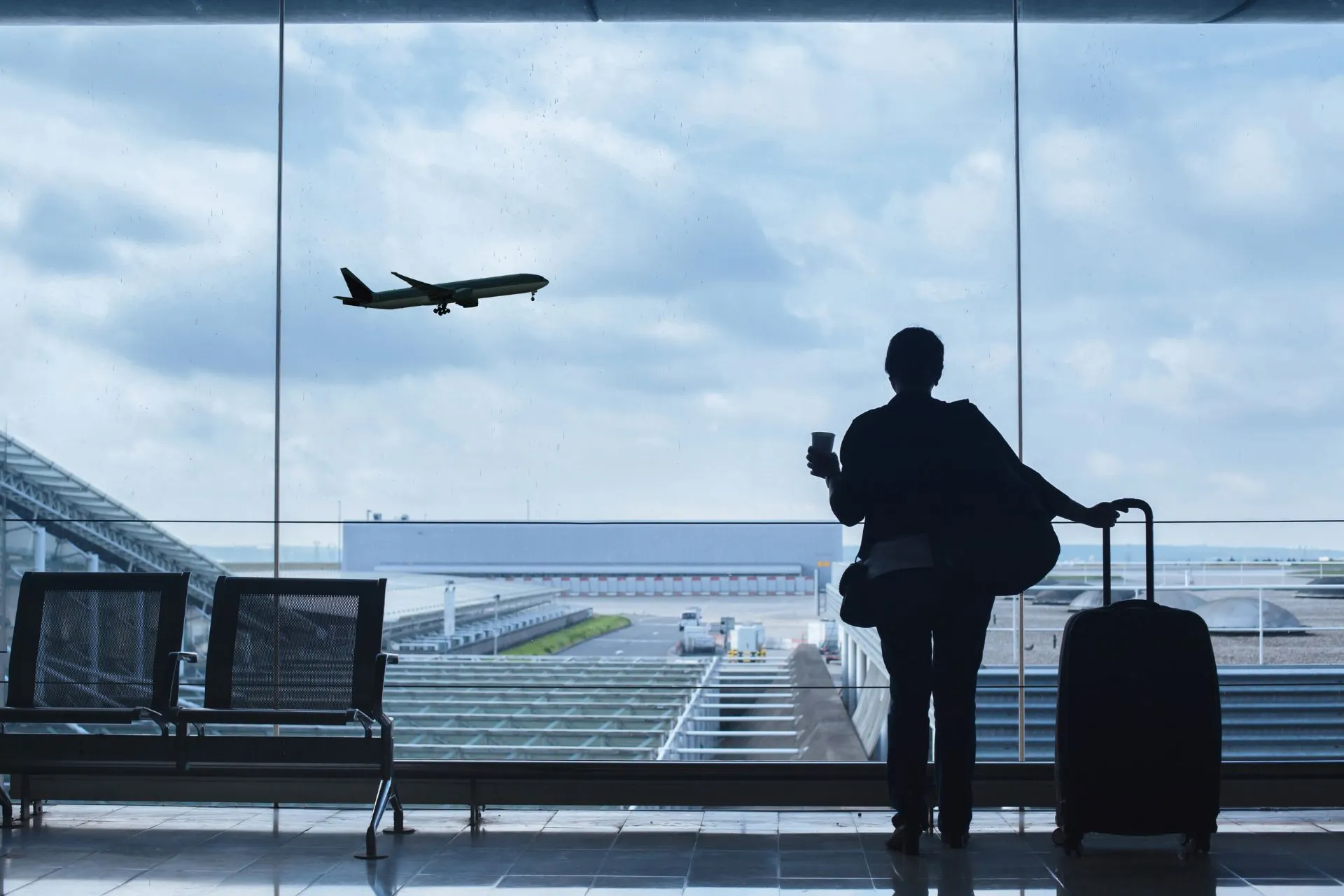 Reiziger met koffers die uitkijkt naar een opstijgend vliegtuig vanuit een groot raam op een luchthaven.