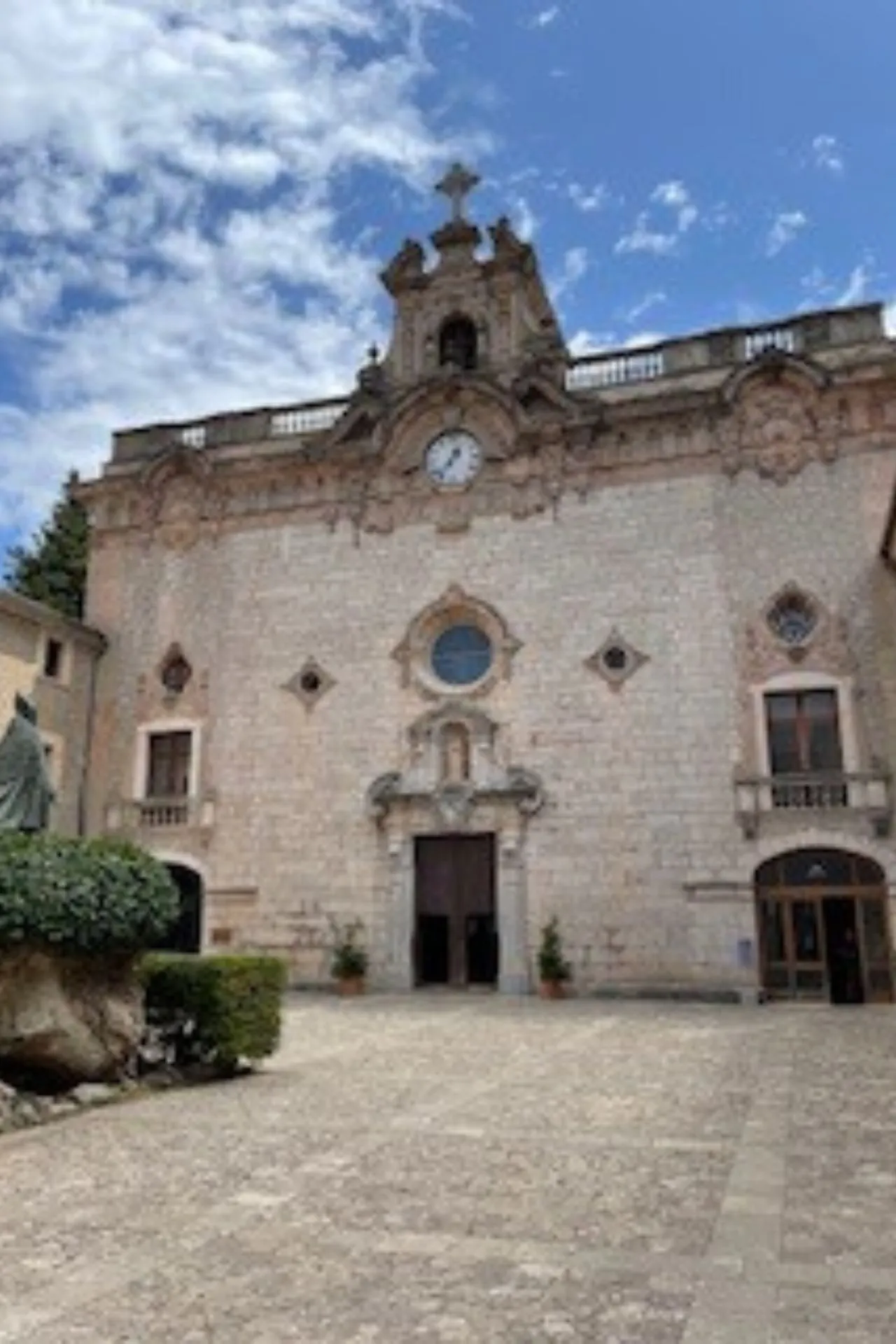 Indrukwekkende historische kerk met gedetailleerde barokke architectuur, grote houten deuren en een ronde glas-in-loodraam boven de ingang. De gevel is gemaakt van lichtgekleurde natuursteen en voorzien van een klok en kruis op de top. Omringd door een rustige geplaveide binnenplaats en groene struiken, onder een heldere blauwe lucht met wolken. Ideaal voor cultuurtoerisme, architectuurliefhebbers en religieuze monumenten op Mallorca.
