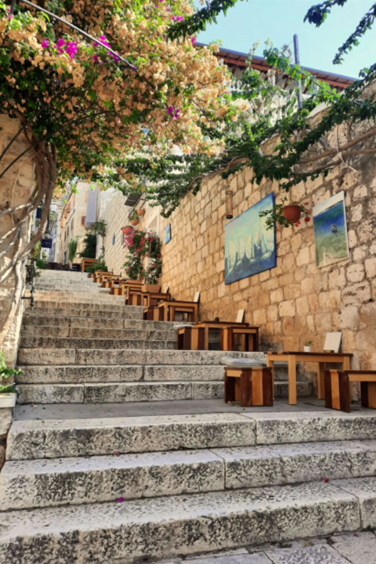 Een charmante stenen trap in Hvar, Kroatië, met houten tafels langs de zijkant, omgeven door bloeiende bougainville en schilderijen aan de muur.