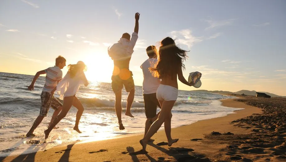 Vijf jongeren springen enthousiast in de lucht op het strand terwijl de zon ondergaat. Hun silhouetten lichten op tegen de kleurrijke lucht en stralen plezier, vrijheid en vriendschap uit.