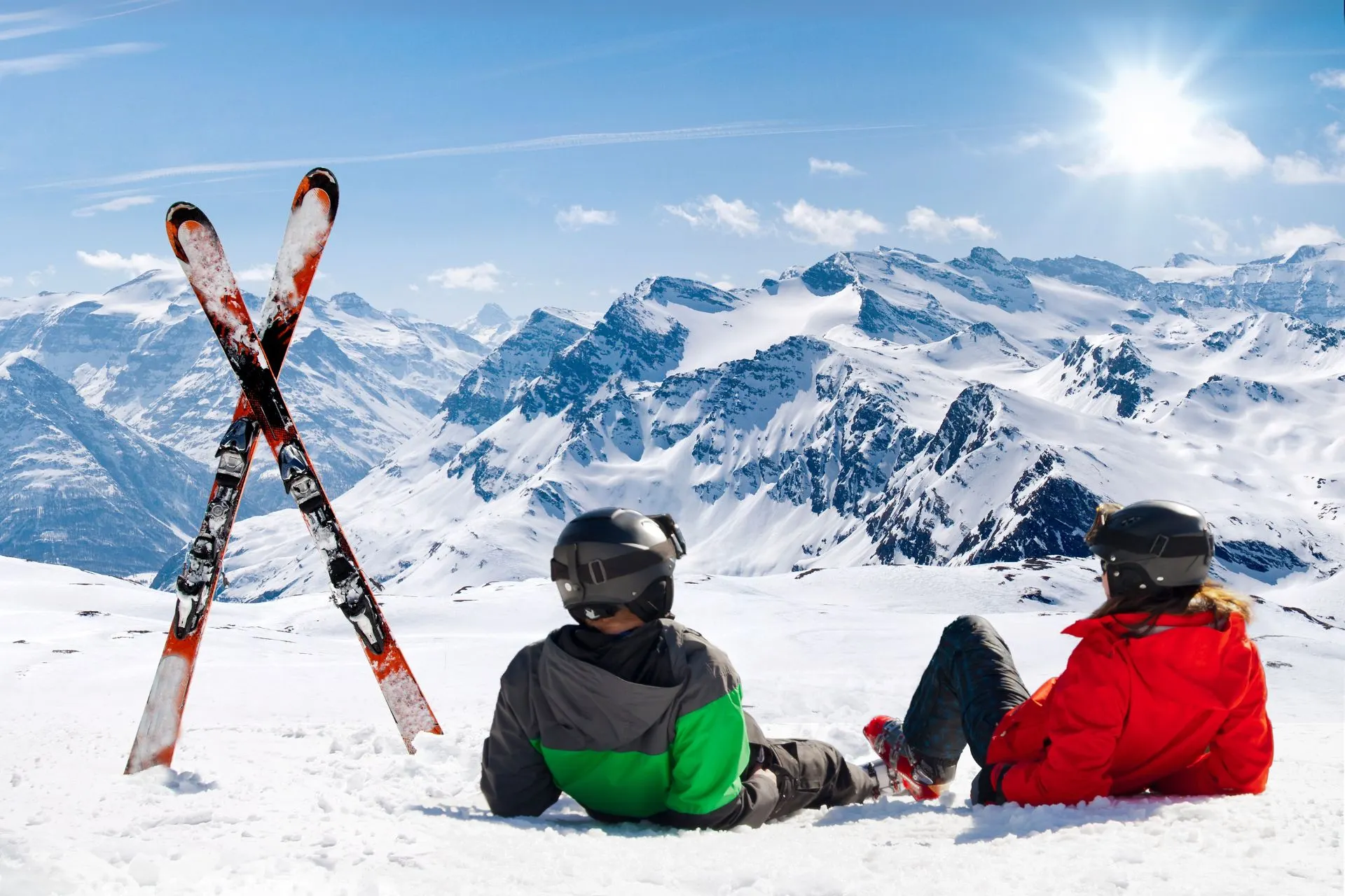 Twee skiërs zitten in de sneeuw met uitzicht op een indrukwekkend berglandschap vol besneeuwde toppen onder een stralende blauwe lucht, met twee gekruiste ski's rechtop in de sneeuw naast hen.