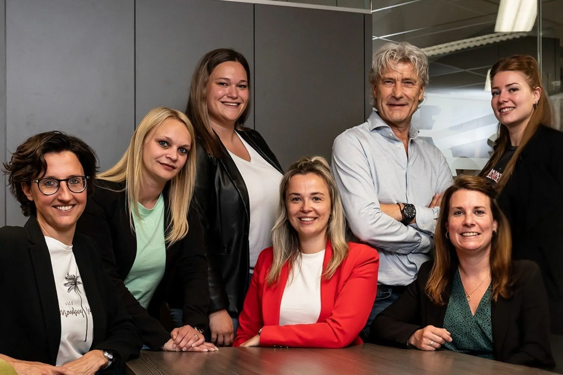 Groepsfoto van een team medewerkers uit Veghel. Zeven collega’s poseren lachend rondom een tafel in een moderne kantooromgeving. De groep bestaat uit vijf vrouwen en één man, allen gekleed in zakelijke, maar informele kleding. Op de achtergrond zijn grijze kasten en een glazen wand zichtbaar.