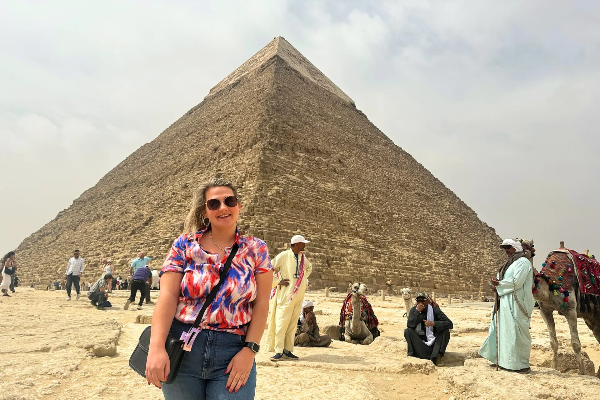 Naomi poseert lachend voor de iconische piramides van Gizeh in Egypte, met zandduinen en een heldere lucht op de achtergrond.