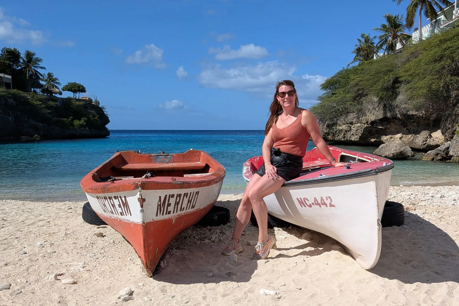 Twee kleurrijke boten staan op een wit zandstrand met helderblauw water en rotsachtige kustlijn op Curaçao. De tropische omgeving met palmbomen en azuurblauwe zee maakt dit een perfecte plek voor een ontspannen vakantie, strandwandelingen en fotografie. Ideaal voor reizigers die authenticiteit en natuur waarderen.