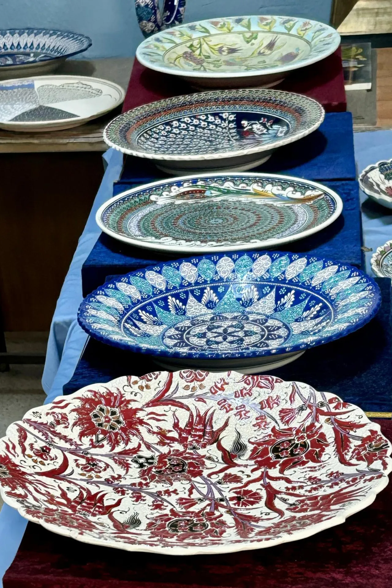 Een collectie handbeschilderde keramische schalen in verschillende kleuren en patronen, tentoongesteld op een tafel in Avanos, Cappadocië, Turkije. Op de voorgrond ligt een grote schaal met rode bloemenmotieven op een witte achtergrond, terwijl andere schalen blauwe, groene en kleurrijke geometrische ontwerpen tonen. Deze kunstwerken weerspiegelen het traditionele Turkse vakmanschap en zijn populair als decoratieve items en souvenirs. Perfect voorbeeld van ambachtelijke keramiek die de rijke cultuur van Anatolië benadrukt.