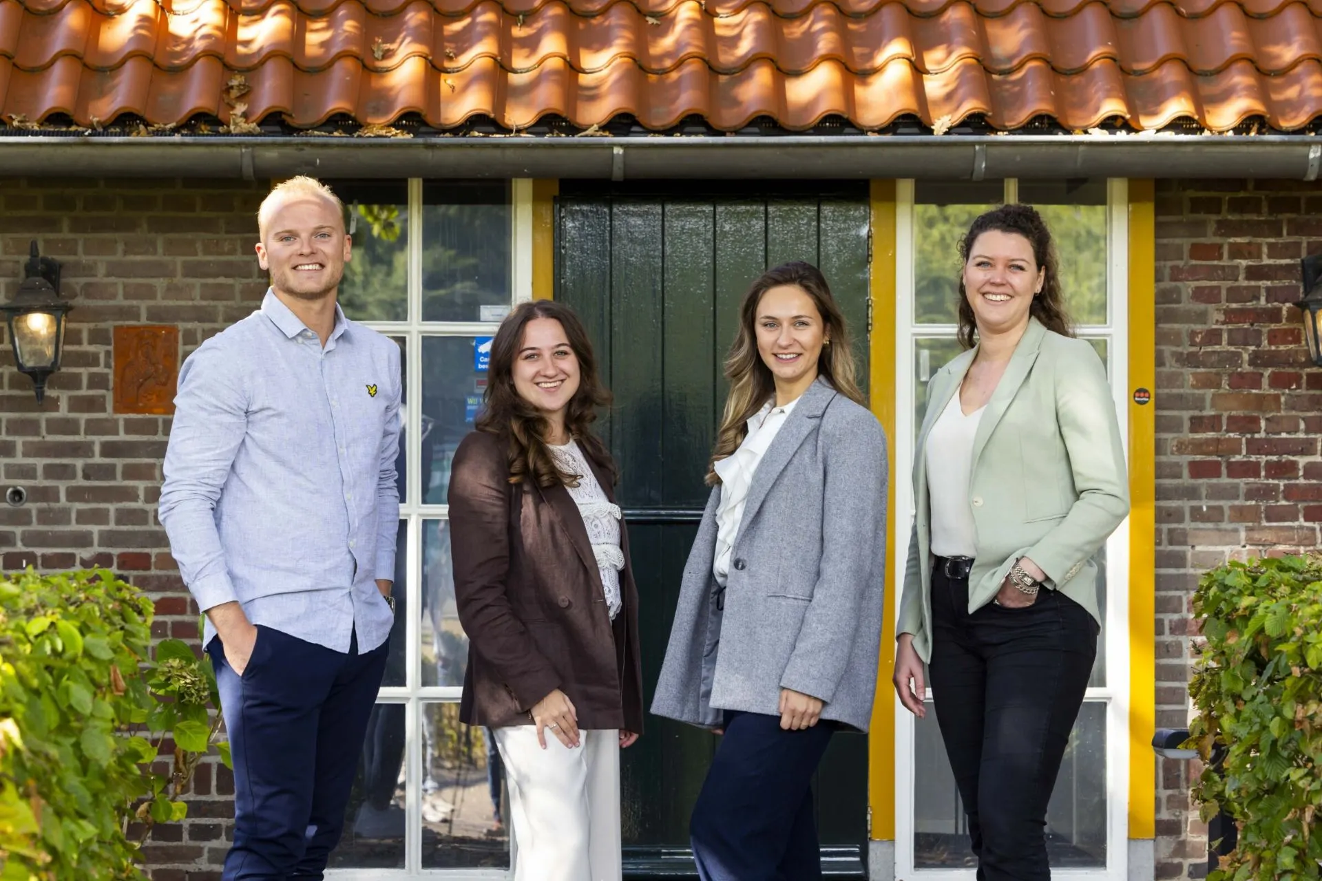 Leonie Wünker van Klooster Reizen, June Römer van Kupers Reisinfo, Emmie de Wit van Travelcenter Nederland en Justin Goverde van Pelikaan Travel Group poseren samen voor een traditionele bakstenen gevel met een zwarte deur en gele kozijnen. Het oranje pannendak en de groene struiken geven de foto een warme, landelijke uitstraling.