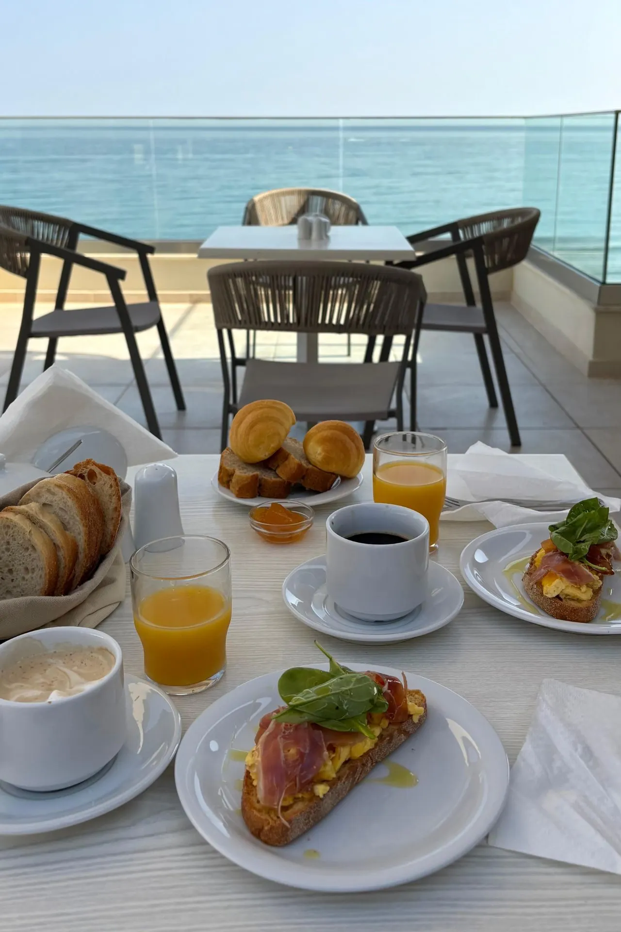 Ontbijttafel met vers brood, croissants, koffie, sinaasappelsap en toast met ham en ei, met uitzicht op de zee vanaf het terras van Batis Beach Hotel.