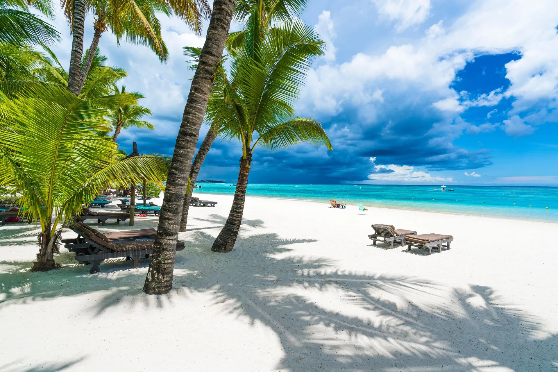  Schaduwrijke palmbomen langs een wit zandstrand met ligstoelen, kijkend naar de azuurblauwe zee en een dreigende, maar prachtige lucht vol wolken.