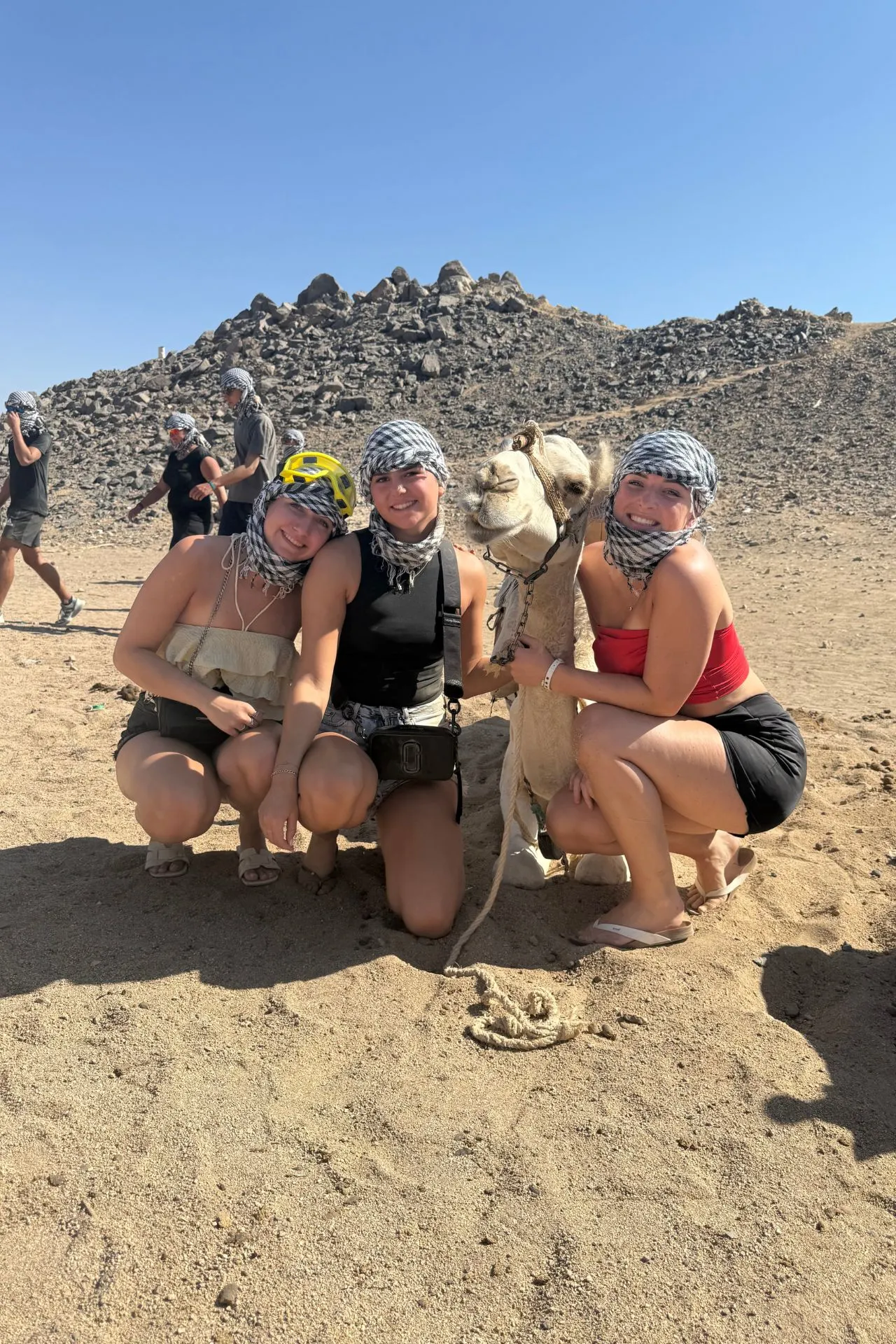 Drie personen gehurkt naast een witte kameel in een woestijnlandschap met rotsachtige heuvels op de achtergrond. Ze dragen sjaals om hun hoofd en lichte zomerkleding. Andere mensen en kamelen zijn zichtbaar in de verte.


