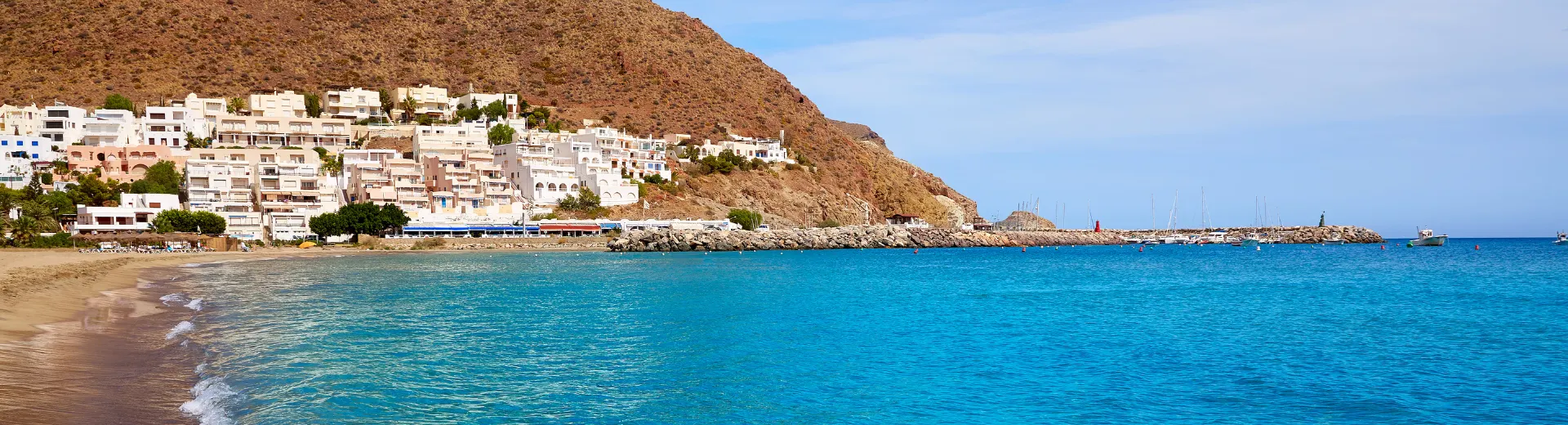 Rustig strand met helderblauw water en een zachte zandkust, gelegen naast een heuvel met witte gebouwen die trapsgewijs tegen de rotswand zijn gebouwd. Op de achtergrond is een kleine haven zichtbaar met enkele boten en een pier, terwijl de lucht helder en licht bewolkt is. Deze schilderachtige kustlocatie is ideaal voor een ontspannen vakantie, strandwandelingen en genieten van een authentieke mediterrane sfeer in een rustige omgeving.