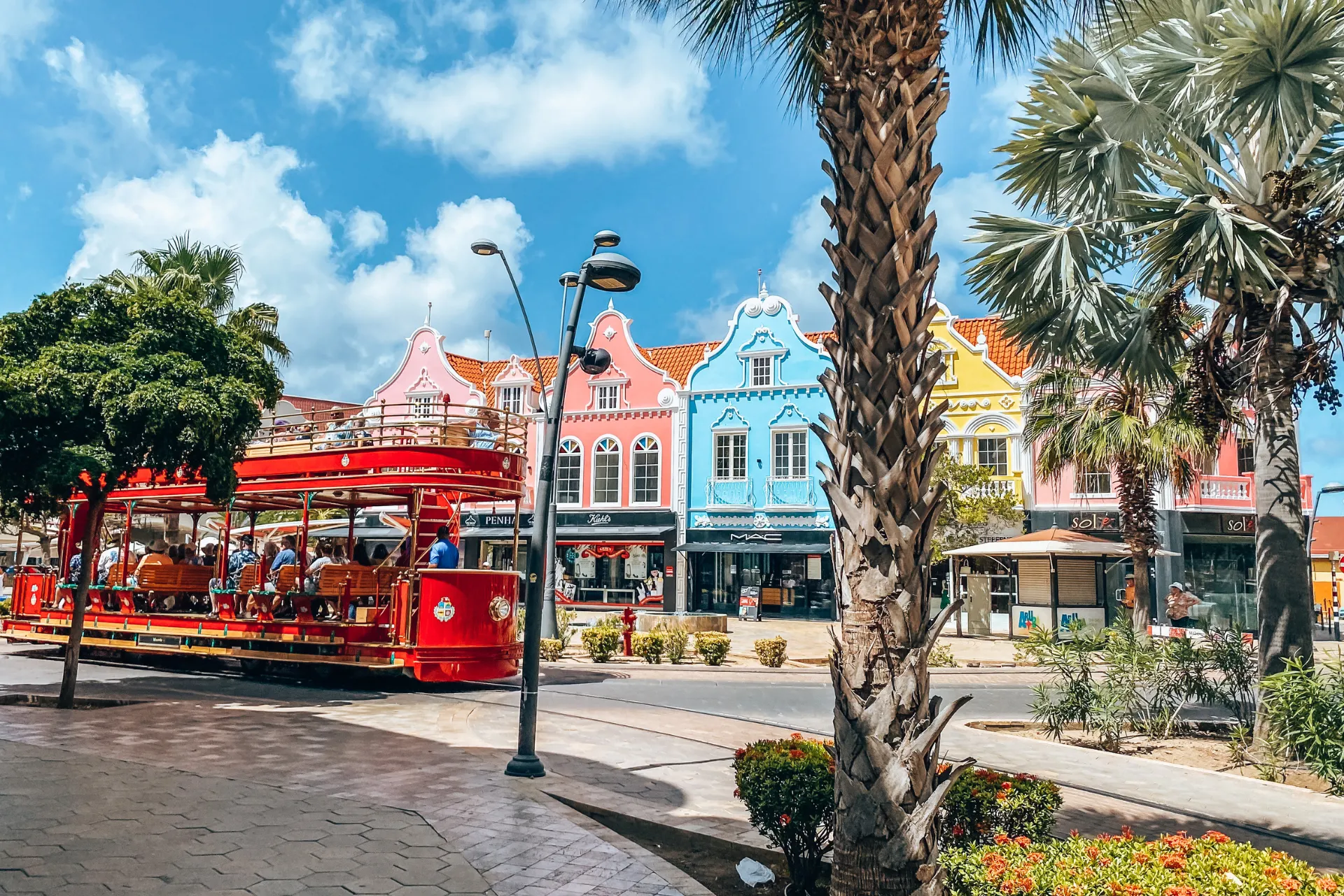 Kleurrijke koloniale gevels in pastelroze, blauw en geel langs een straat in Oranjestad, Aruba, met een rode open toeristentram op de voorgrond, omringd door palmbomen en een heldere blauwe lucht, een populaire bezienswaardigheid voor eilandbezoekers.