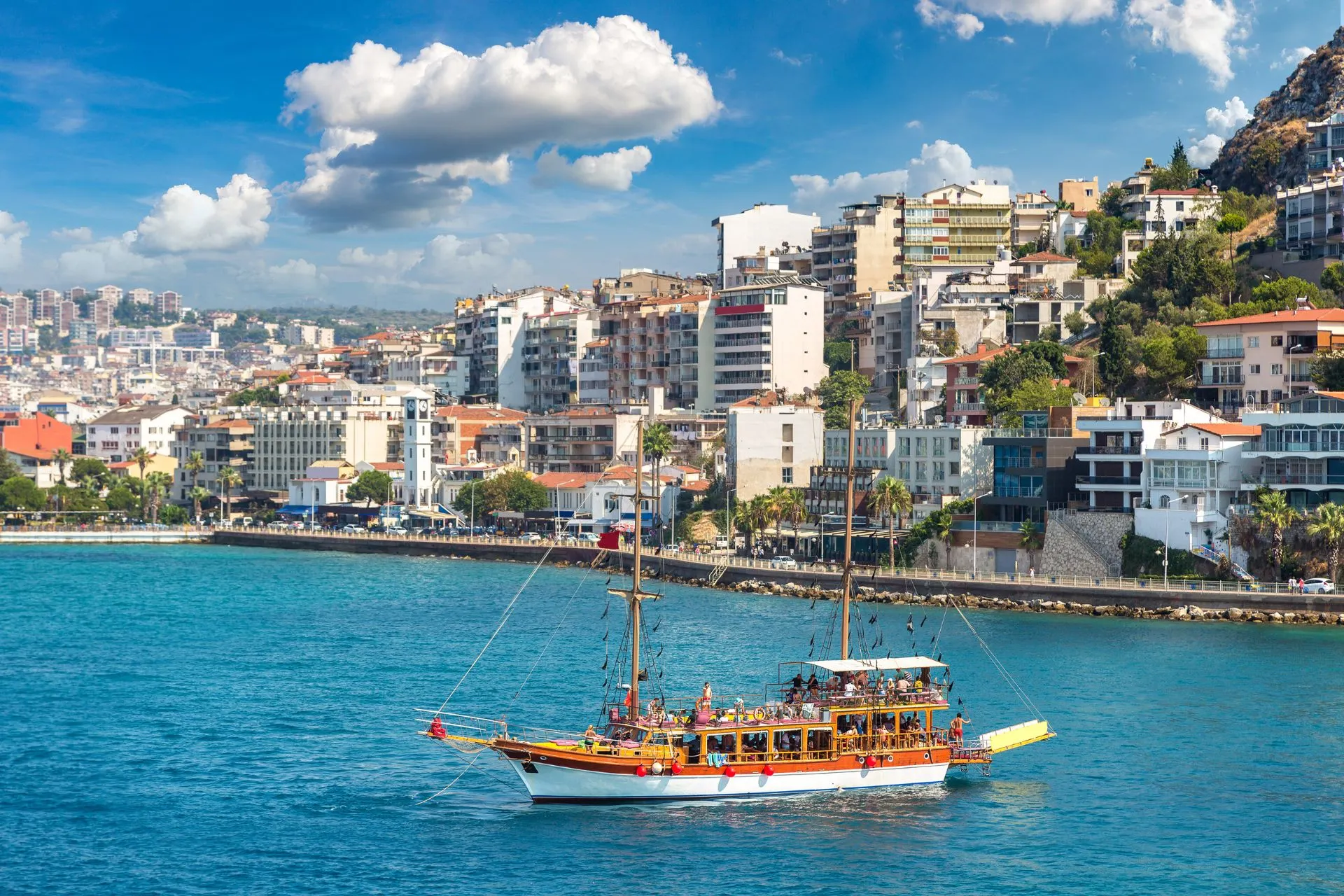 Kleurrijke houten boot vol toeristen vaart langs de kust van Kuşadası met de stad op de achtergrond.