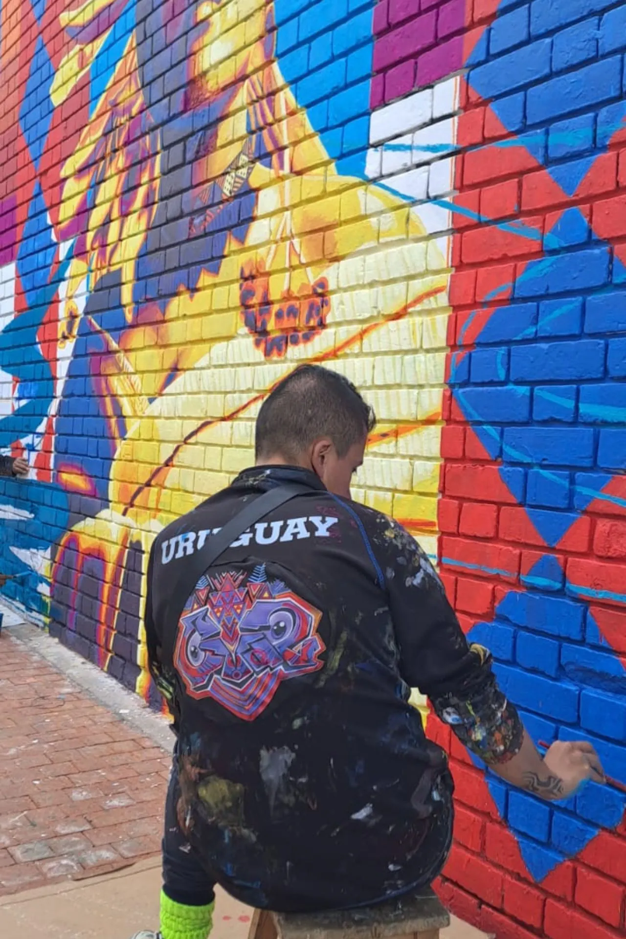 Straatkunstenaar schildert een kleurrijke muurschildering op een bakstenen muur, met geometrische patronen en een figuur in gele tinten.