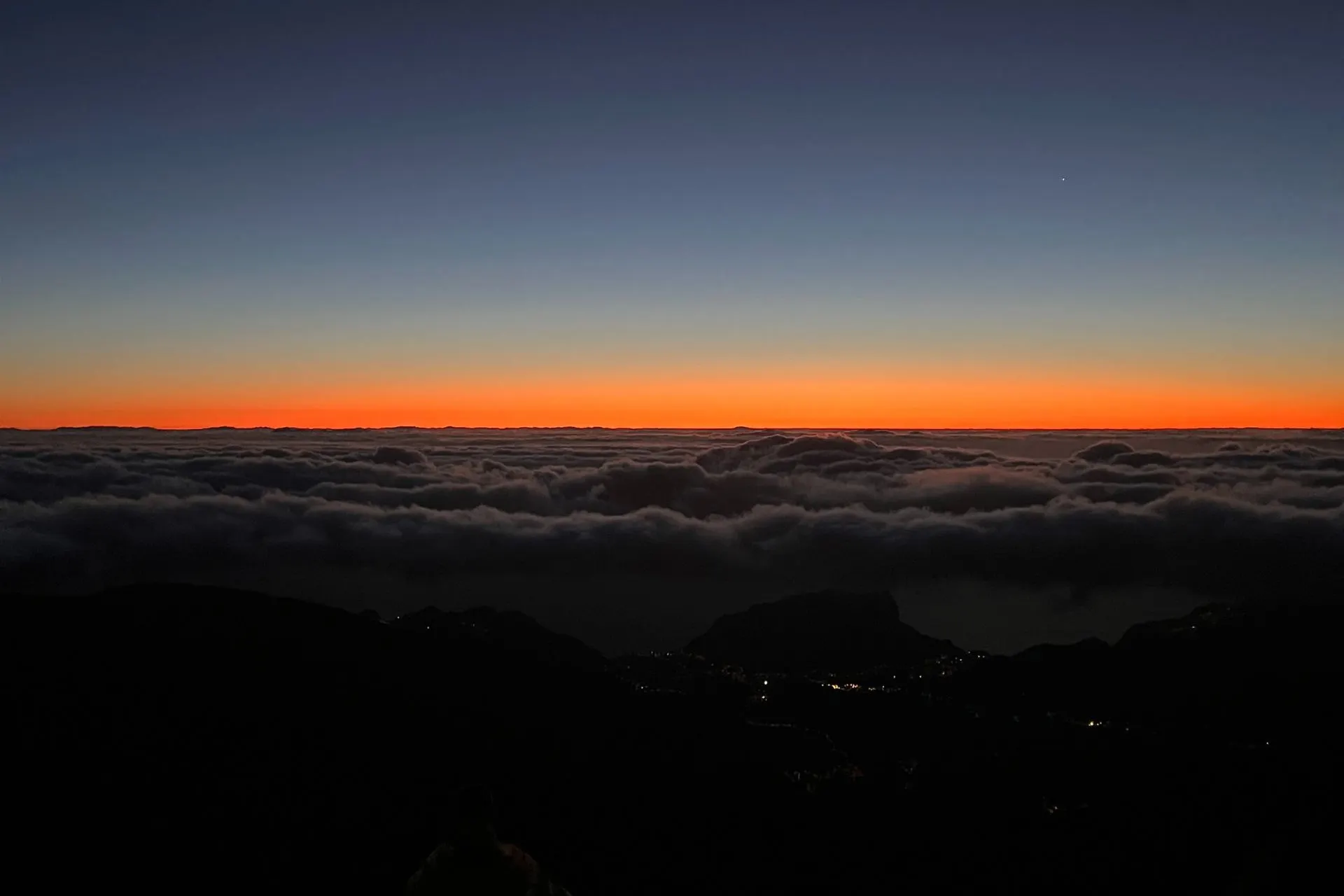 Donkere bergsilhouetten onder een heldere hemel bij zonsopgang op Pico do Arieiro. Een smalle oranje gloed markeert de horizon, terwijl de lucht overgaat van diepblauw naar zwart. Rechtsboven is een halve maan zichtbaar.
