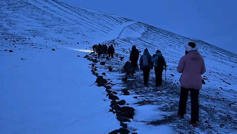 Groep wandelaars op weg naar de top van de besneeuwde Hverfjall‑krater tijdens een winterhike in Noord‑IJsland.
