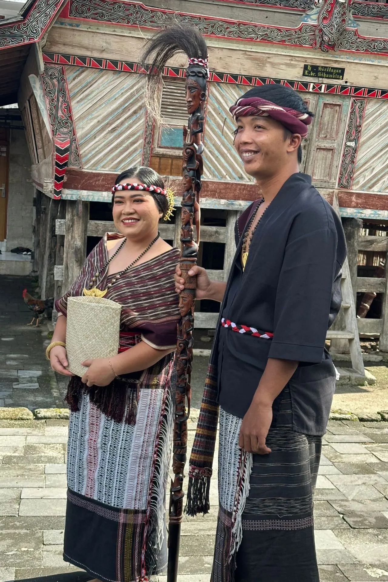Een man en een vrouw in traditionele Batak-kleding poseren glimlachend voor een traditioneel Batak-huis.