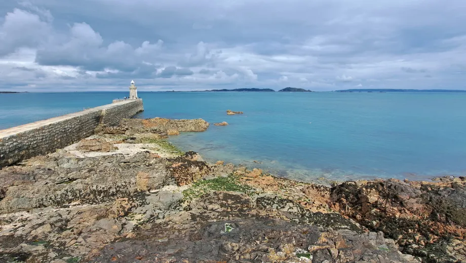 Rotsachtige kustlijn met een lange stenen pier die uitloopt naar een witte vuurtoren aan het einde, omringd door helderblauw zeewater. De lucht is bewolkt, wat zorgt voor een dramatische en contrasterende sfeer boven de oceaan. Op de voorgrond zijn ruige rotsformaties zichtbaar, bedekt met mos en zeewier. Deze schilderachtige locatie is ideaal voor kustwandelingen, fotografie en het ontdekken van maritieme bezienswaardigheden. Perfect voor toeristen die op zoek zijn naar rust, natuur en panoramische uitzichten.
