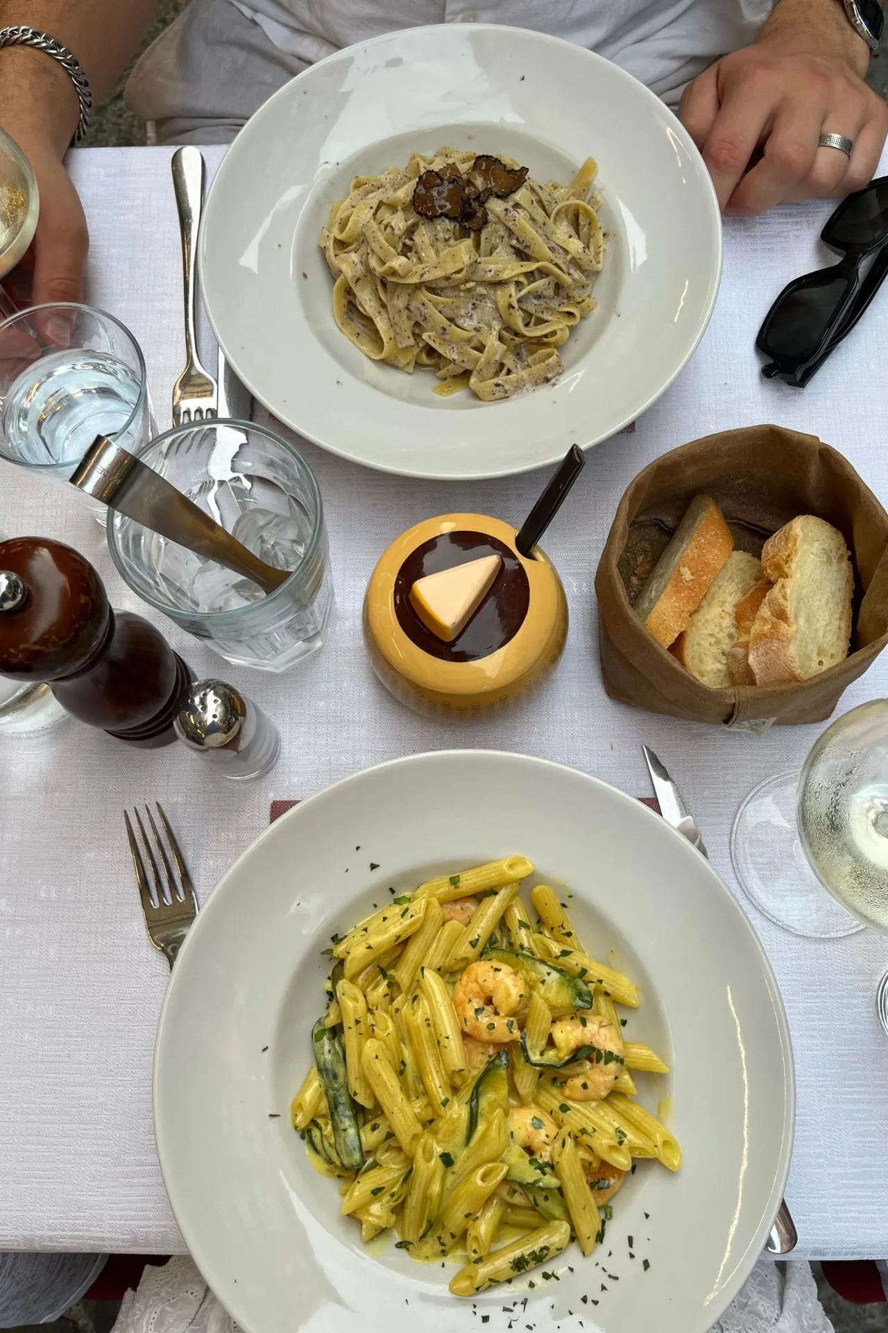 Bovenzicht van een Italiaans diner op een wit tafellaken, met een bord tagliatelle met truffel en roomsaus, een bord penne met garnalen en courgette in saffraansaus, vers brood, glazen water en witte wijn.