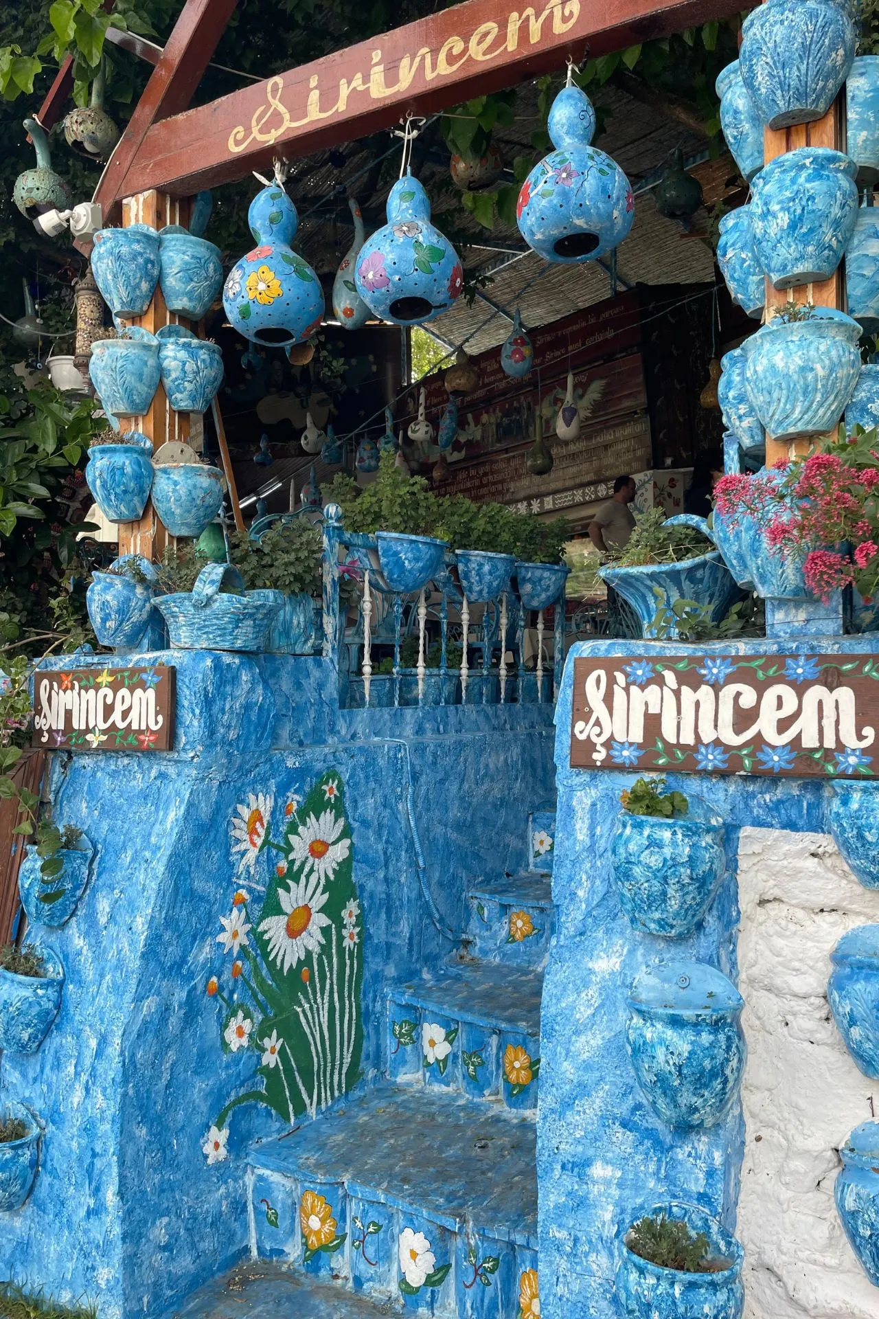 Kleurrijke ingang van een winkel of atelier in Şirince met blauwe handbeschilderde bloempotten, decoraties en het bord ‘Şirincem’.