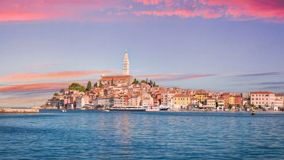 "Schitterend uitzicht op Rovinj in Istrië, Kroatië, met kleurrijke historische huizen langs de kust en de iconische Sint-Eufemiakerk op de heuveltop. De roze en paarse tinten van de zonsondergang weerspiegelen in het kalme water van de Adriatische Zee. Rovinj is een populaire vakantiebestemming in Istrië, bekend om zijn pittoreske straatjes, mediterrane sfeer, stranden en cultuur. Ontdek Kroatische charme en gastronomie."