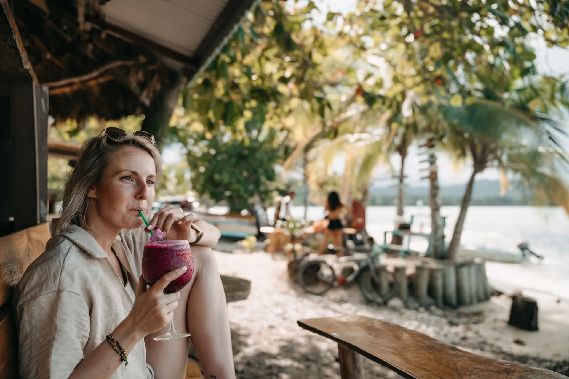 Een vrouw drinkt een kleurrijke smoothie op een strand met een tropische achtergrond vol palmbomen.