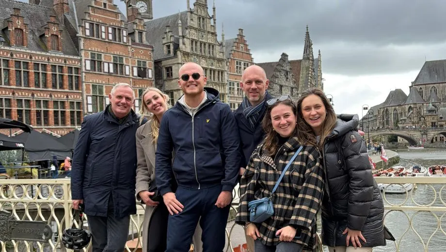 Groepsfoto van reisspecialisten van DTA op een brug in Gent, met historische gevelhuizen, een kerk en de rivier de Leie op de achtergrond.