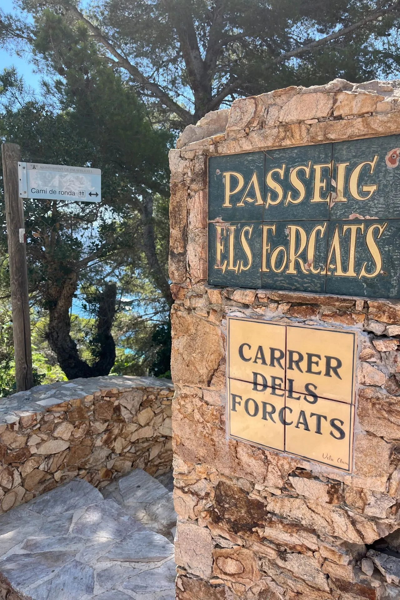 Stenen muur met straatnaamborden ‘Carrer dels Forcats’ en ‘Passeig Els Forcats’, omringd door bomen en een wandelpad.