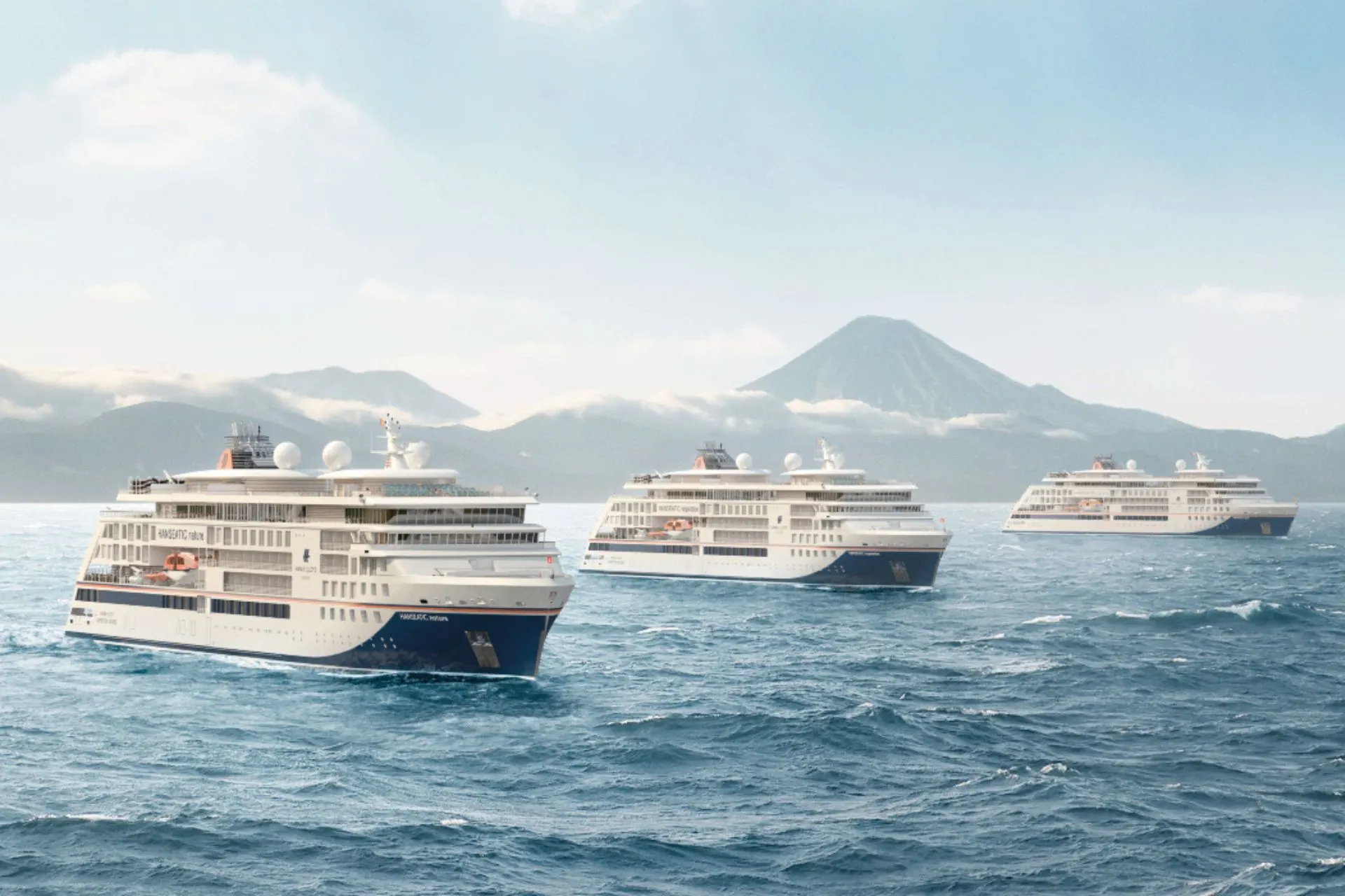 De drie moderne expeditieschepen Hanseatic nature, Hanseatic inspiration en Hanseatic spirit van Hapag-Lloyd varen samen op open zee met een vulkanisch eiland en wolkenflarden op de achtergrond.